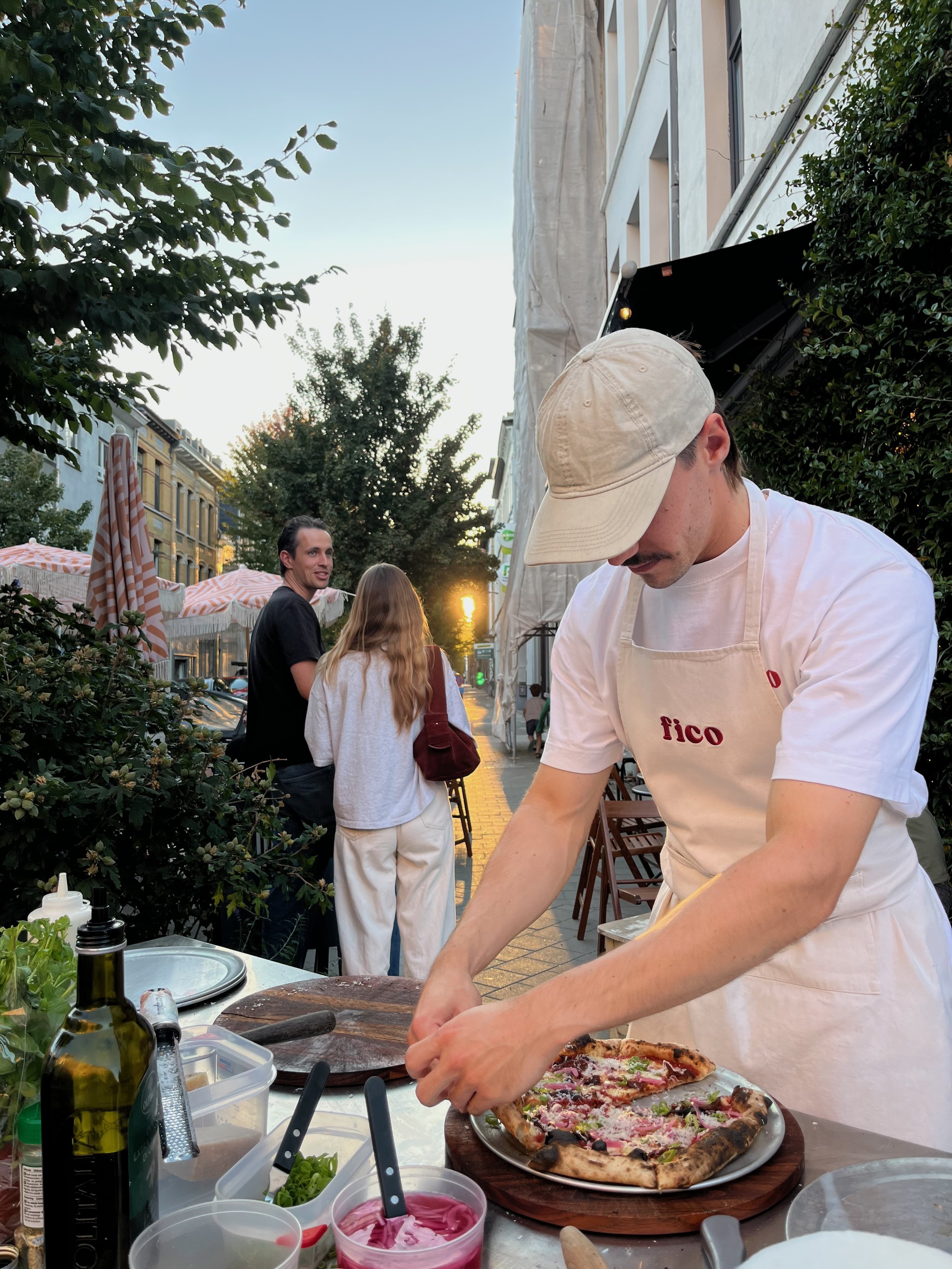 Een persoon is een pizza aan het klaarmaken op een terras in de ondergaande zon. Op de achtergrond staan bomen en parasols en er zijn mensen gezellig aan het babbelen.