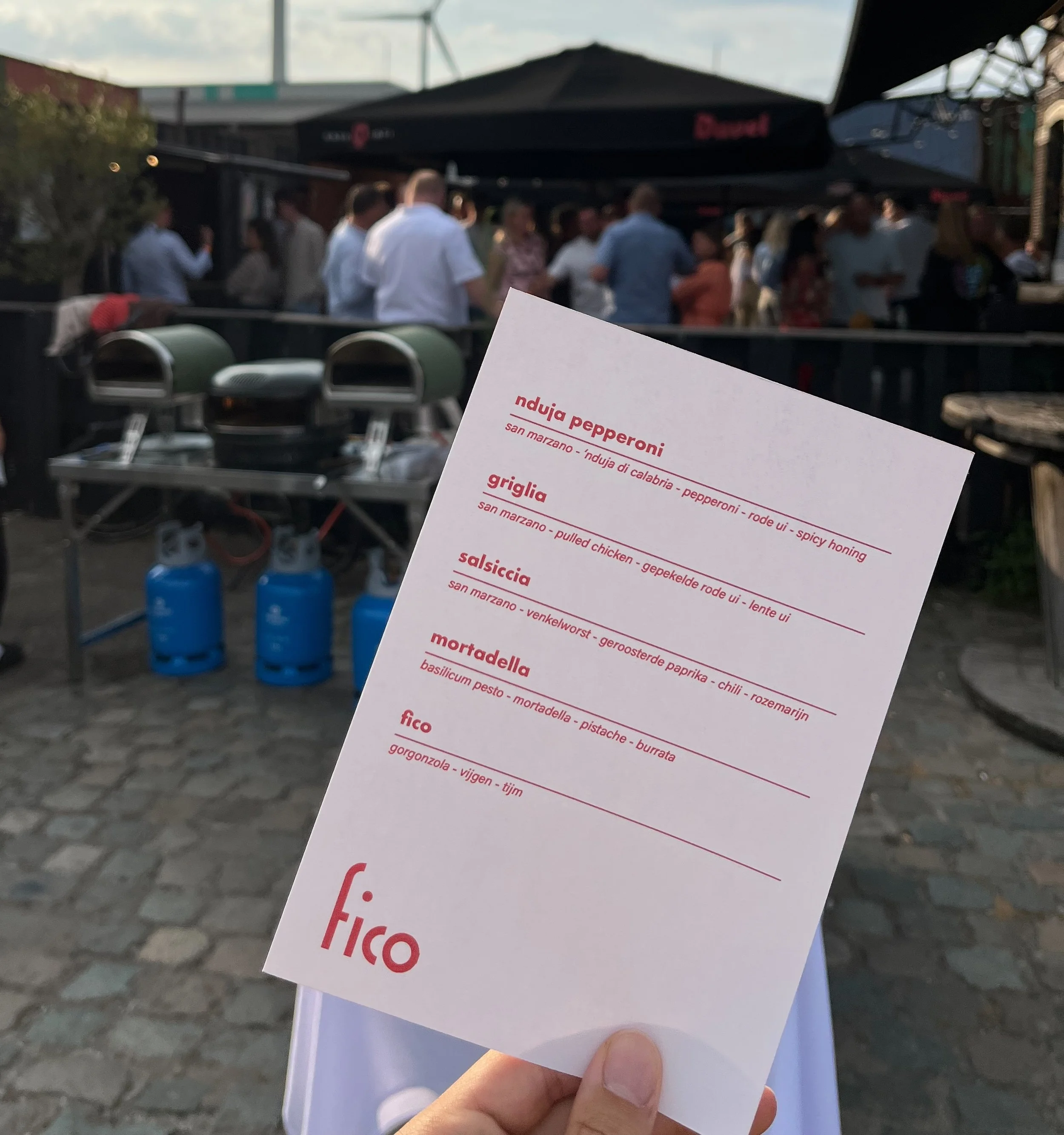 Een hand die een wit menu met rode tekst vasthoudt. In de wazige achtergrond zijn er mensen aan het socializen en plezier aan het maken op een verjaardagsfeest buiten.