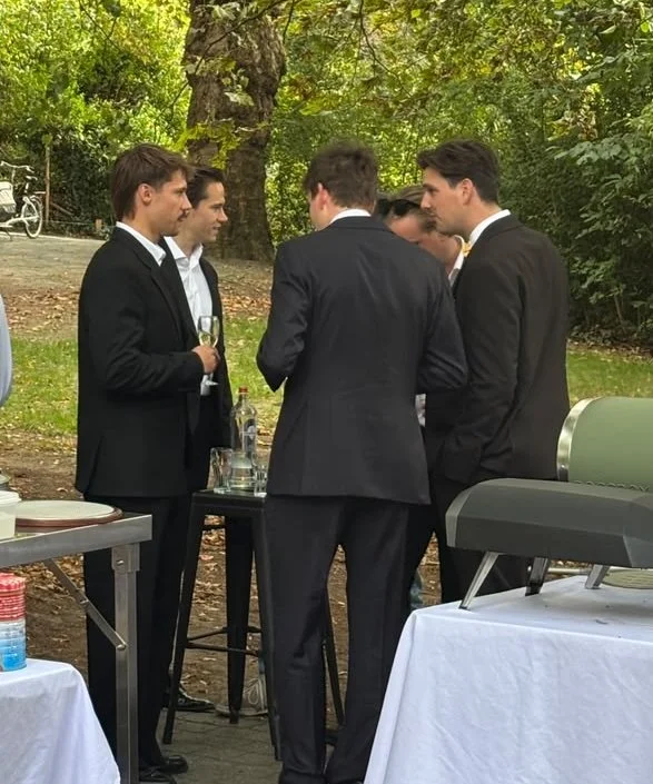 Een aantal mannen in een chique kostuum staan op een trouwfeest rond een tafel met een glas in de hand naast de pizza ovens.