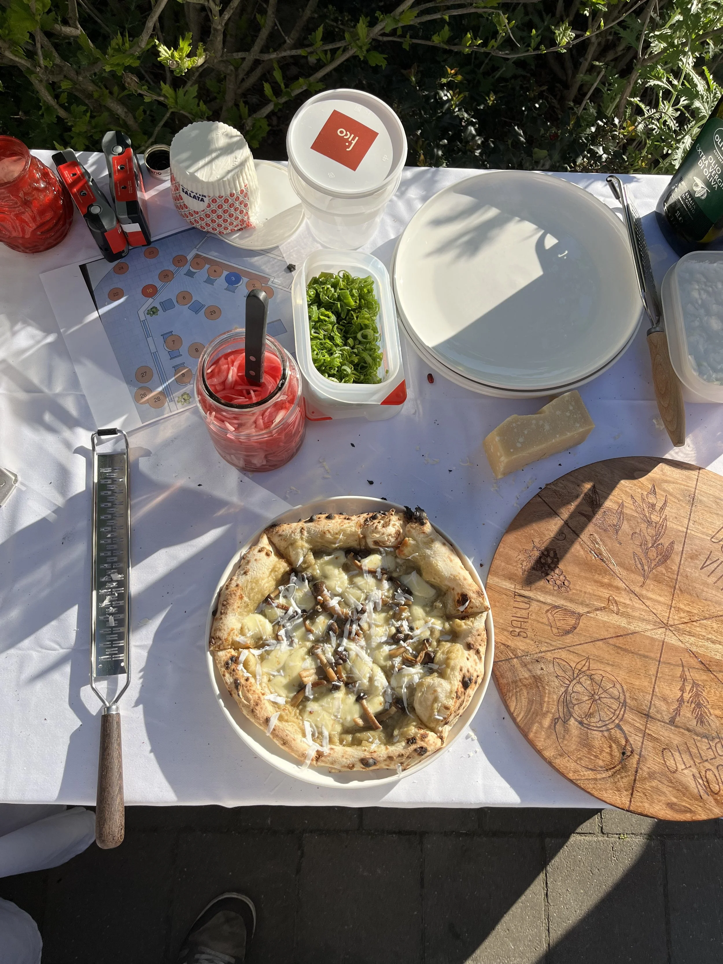 Outdoor table with pizza topped with cheese and mushrooms, a container of sliced green onions, a cheese block, plates, a jar of pickled vegetables, a jar of sliced red vegetables, paper napkins, and various utensils on a white tablecloth.