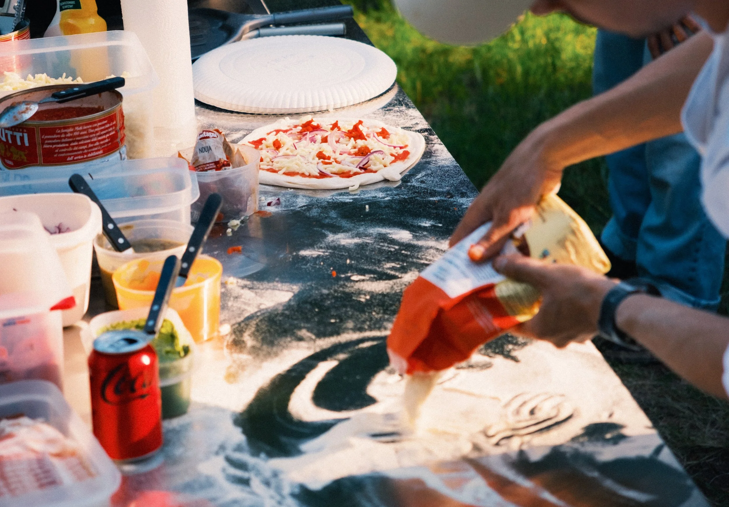 Een persoon die bloem op een oppervlak giet om er pizza op te maken. Er staan verschillende ingrediënten op tafel en er ligt een pizza klaar om te bakken.