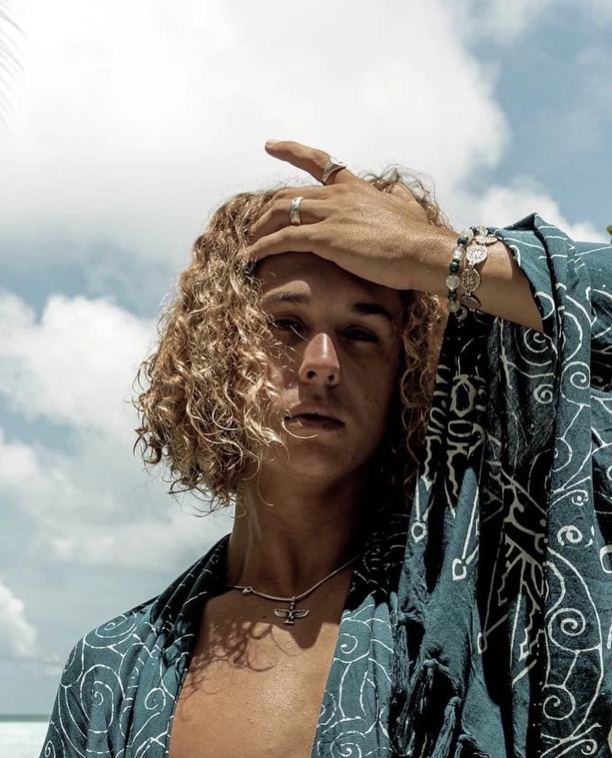 A woman with curly blonde hair and light skin standing outdoors against a cloudy sky. She is wearing a patterned shirt, jewelry including rings, bracelets, a necklace, and has a serious expression with her hand on her forehead.