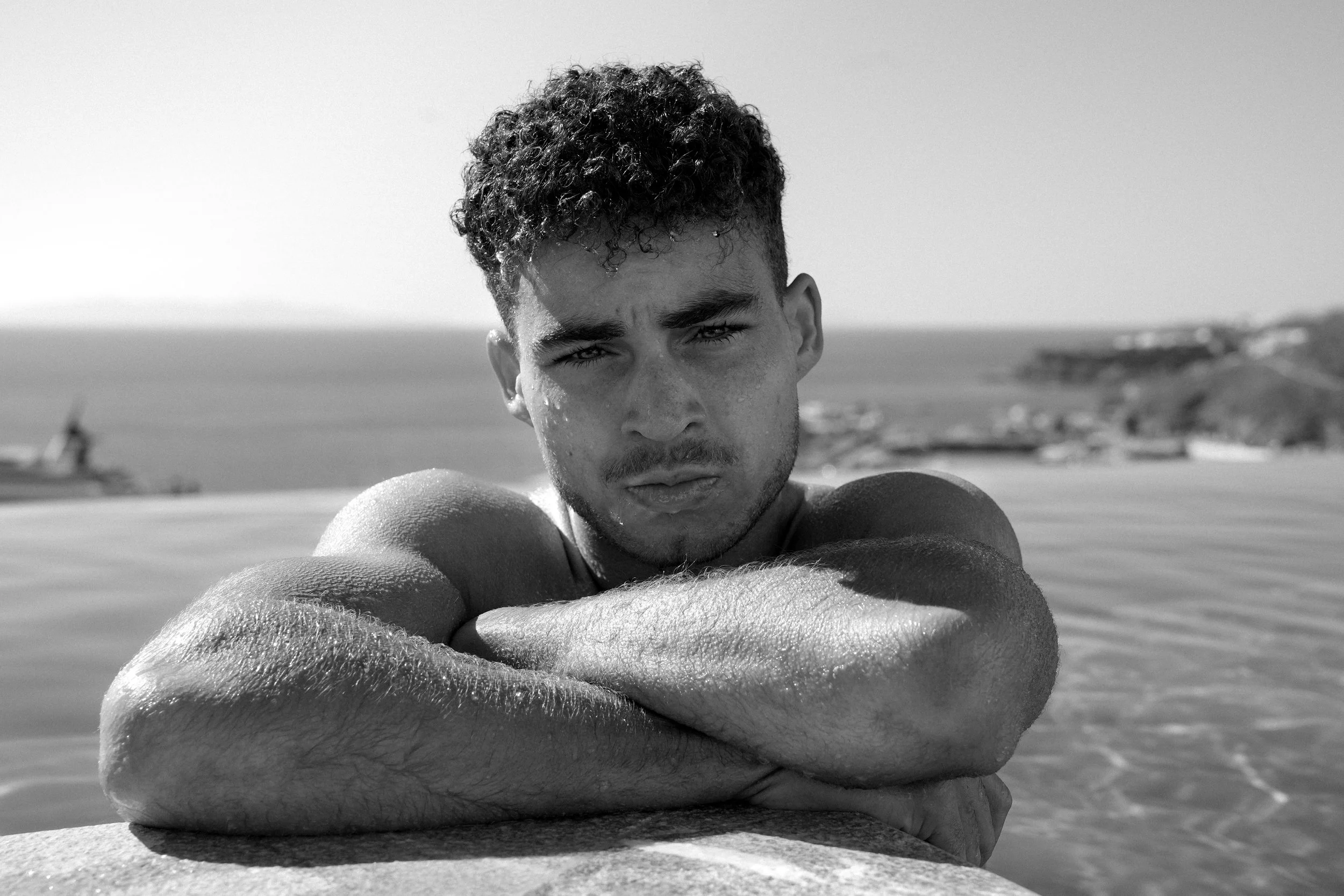 Black and white photo of a young man with curly hair resting his arms on a ledge at the beach, looking directly at the camera with a serious expression, with water and distant land in the background.