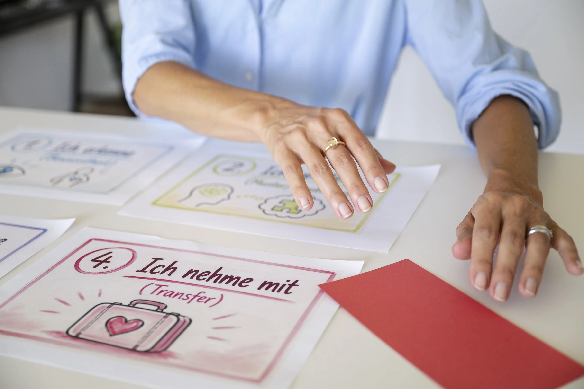 Eine Person zeigt mit einem Finger auf eine handschriftlich gestaltete Karte mit der Nummer 4, die den Text 'Ich nehme mit Transfer' enthält, auf einem weissen Tisch.