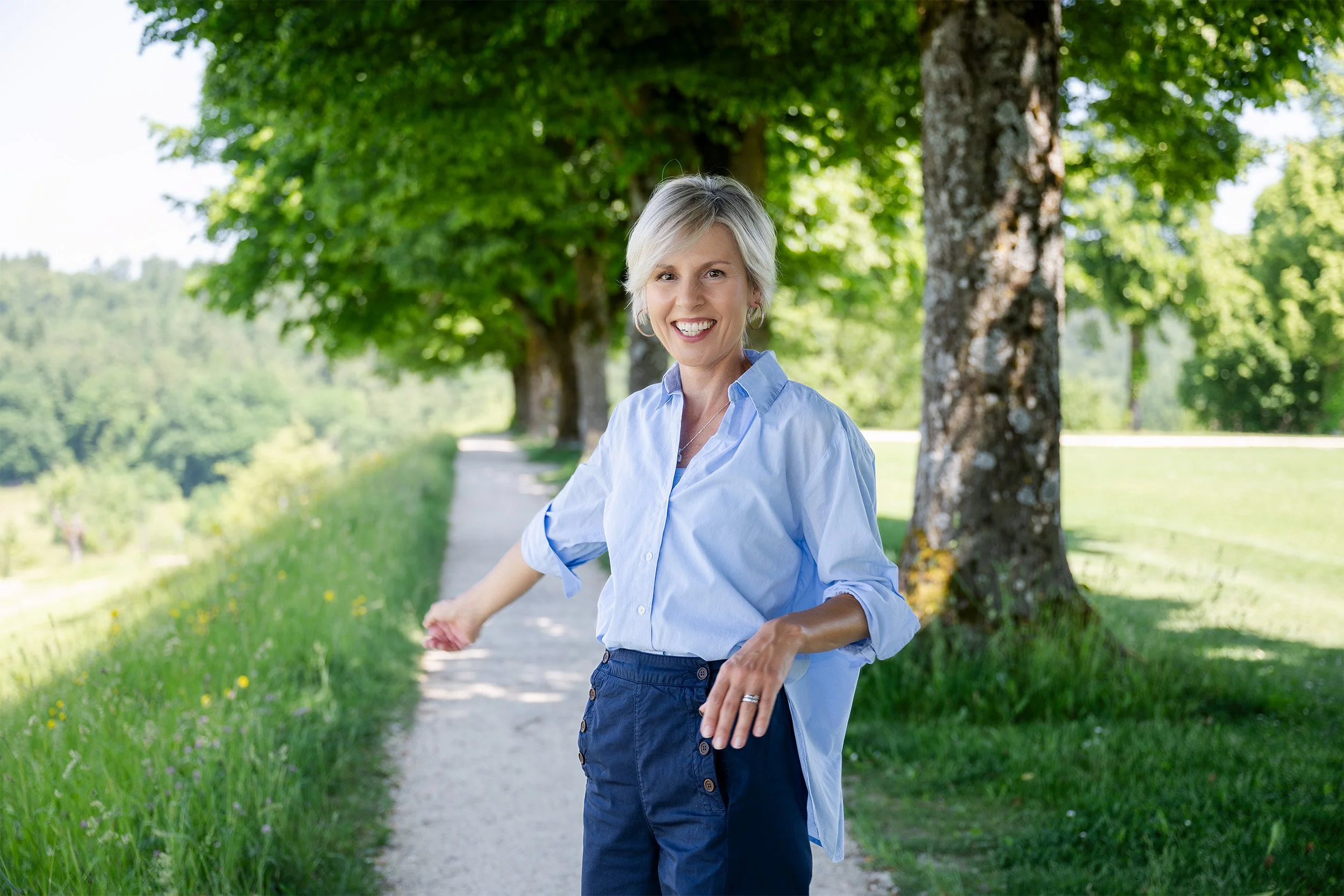 Eine lachende Frau - Sandra Knechtli - mit kurzem Haar in einem hellblauen Hemd und dunklen Hosen steht an einem Weg in einem grünen Park, umgeben von Bäumen und Wiesen.