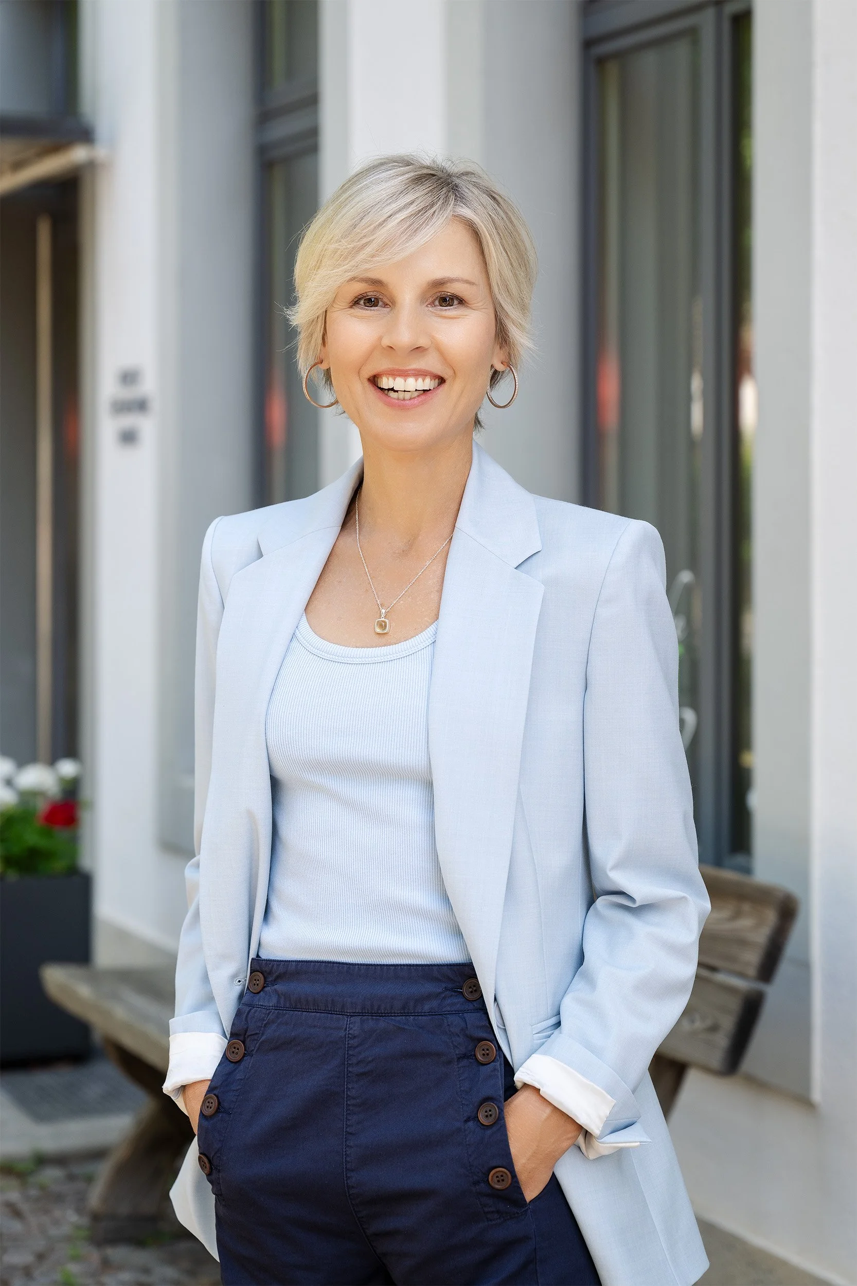 Eine Frau - Sandra Knechtli - mit kurzen blondem Haar, in einem weißen Blazer und blauer Hose, lächelnd vor einem modernen Gebäude mit großen Fenstern.
