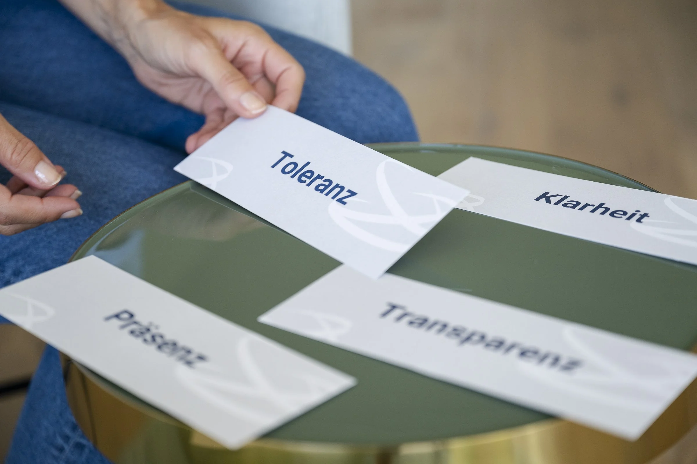 Eine Hand legt eine weisse Karte mit dem Wort "Toleranz" auf einen grünen runden Tisch. Weitere Karten auf dem Tisch tragen Wörter wie "Klarheit" und "Präsenz".