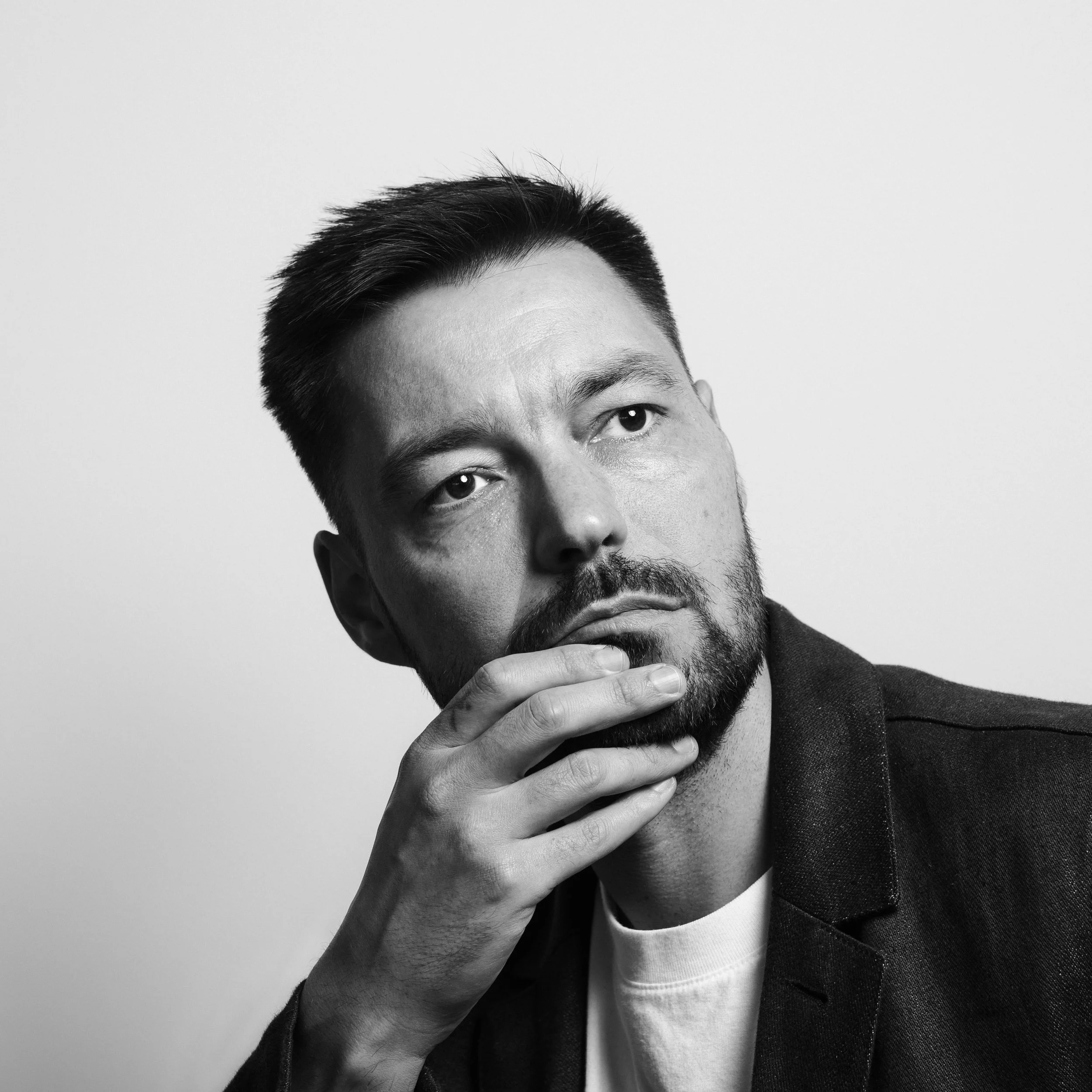 A black and white close-up photograph of a man with short dark hair, a beard, and mustache, wearing a suit jacket over a white shirt. The man is resting his chin on his hand, looking slightly off to the side with a thoughtful expression.