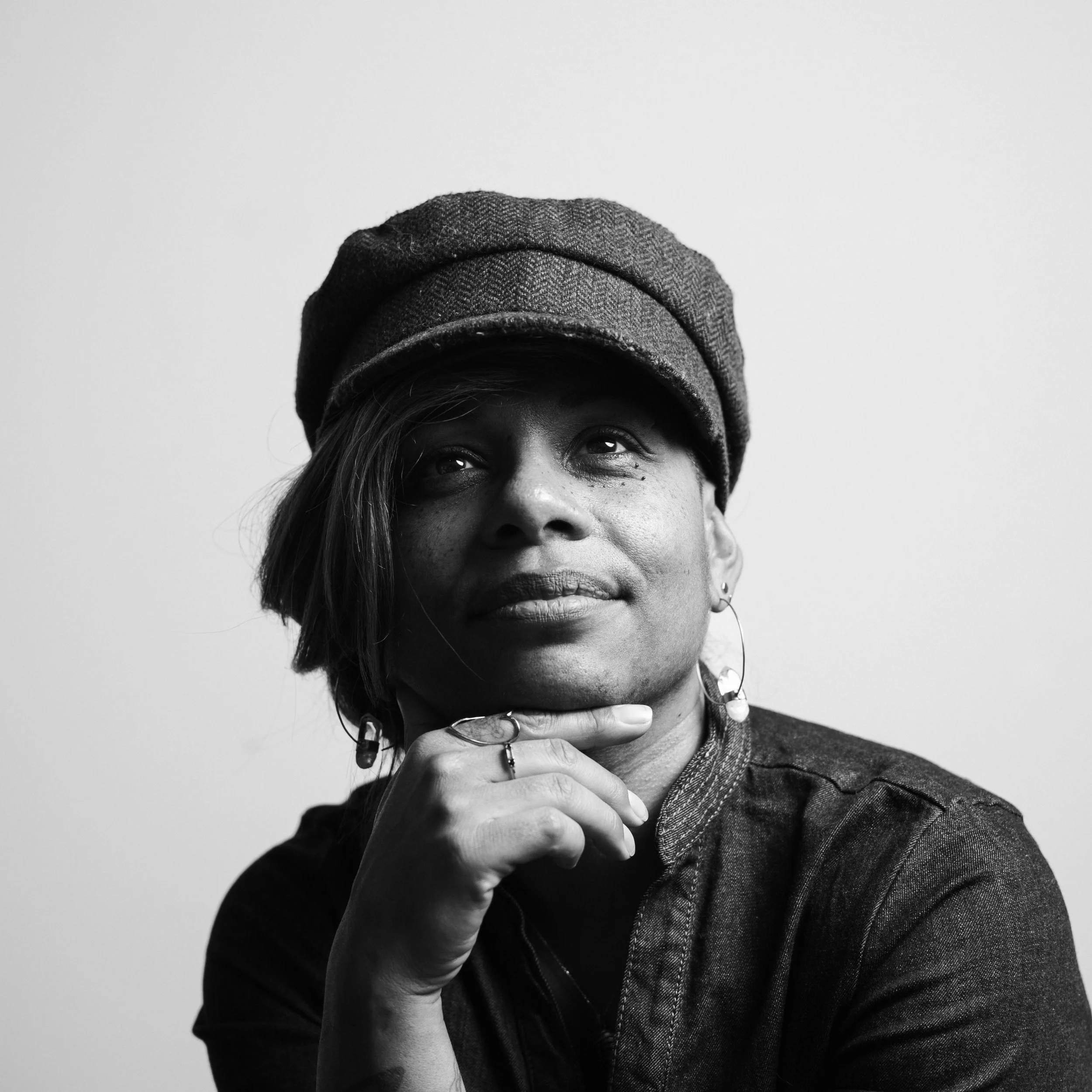 A close-up black and white photo of a woman wearing a wool cap, with her hand resting on her chin, smiling slightly.
