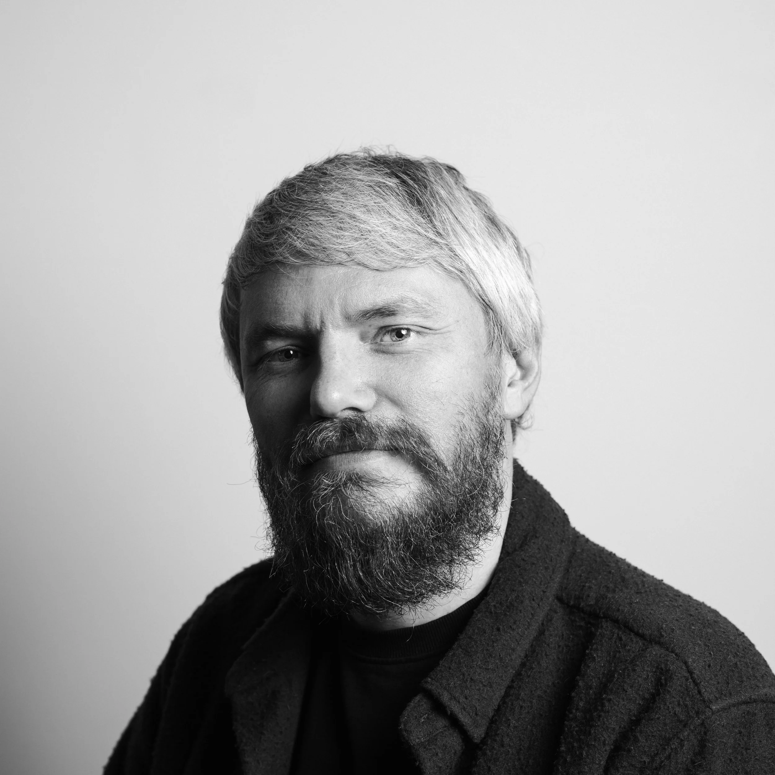 Close-up black-and-white photo of a middle-aged man with short gray hair, a beard, wearing a dark sweater, looking directly at the camera with a serious expression.