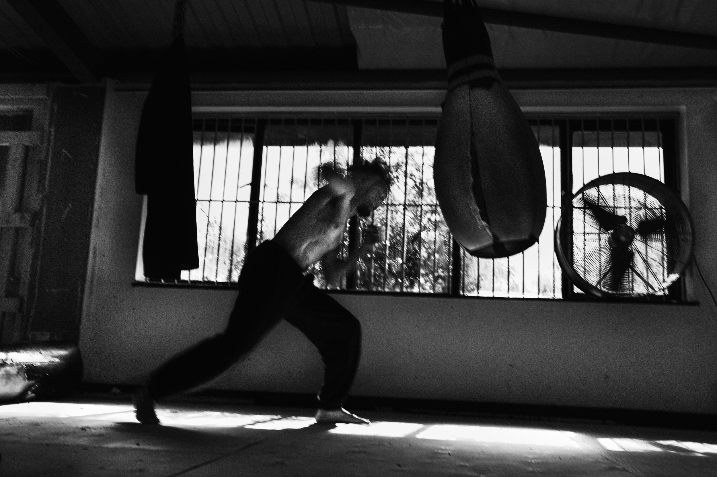 A person practicing yoga or dance indoors next to a large window with metal bars, with a fan visible on the right side. The person is in a dynamic pose with one foot on the ground and the other leg extended behind, and their arms outstretched.