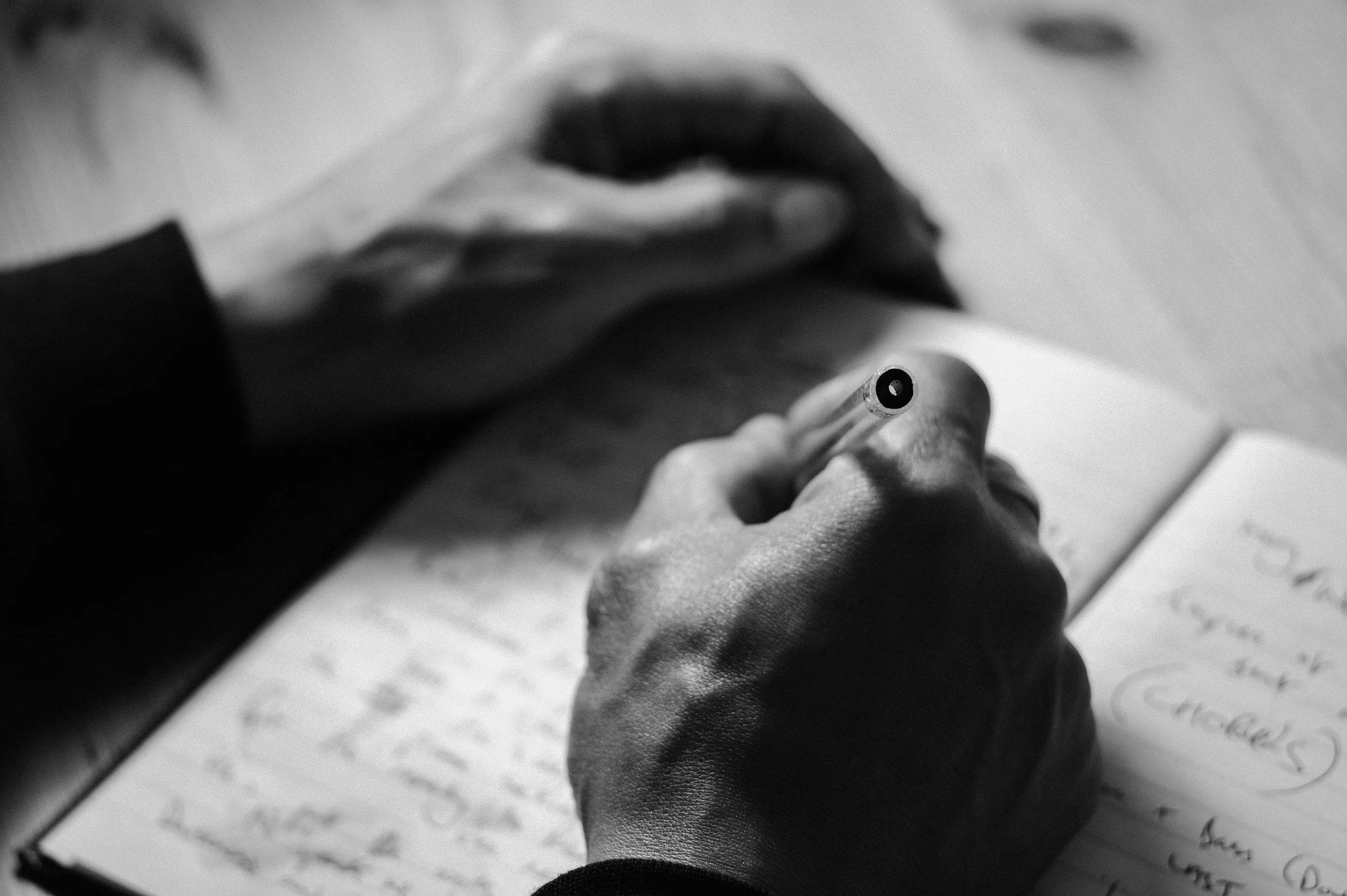 Black-and-white close-up photo of a person's hand writing on a notebook with cursive writing, holding a pen.