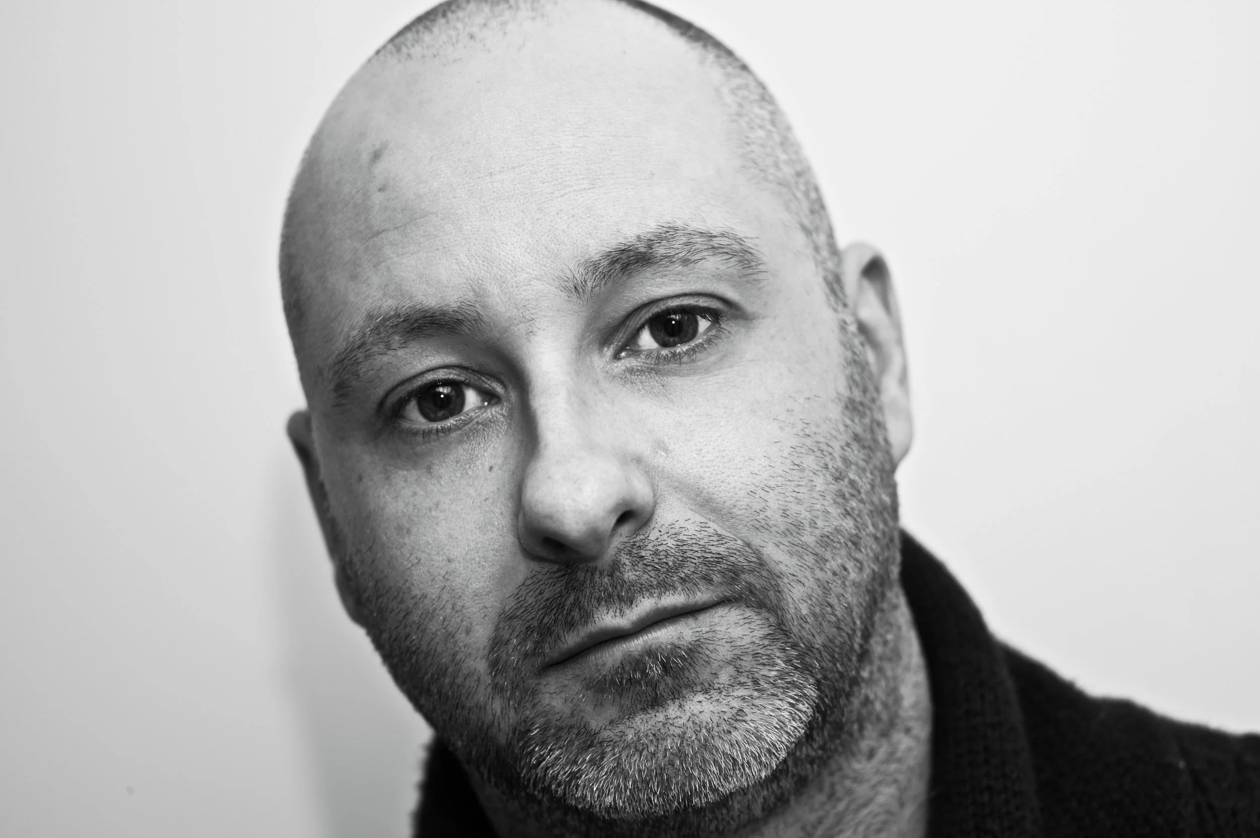 A close-up black and white portrait of a man with one eye partially visible, bald head, subtle facial hair, and wearing a dark top against a plain background.