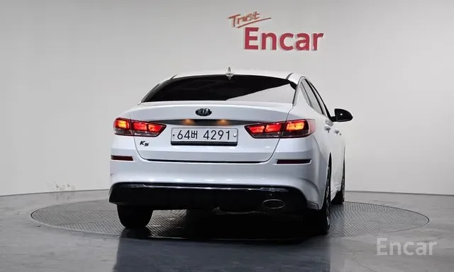 White Kia K5 sedan with visible license plate, parked indoors in front of a white wall with the words 'Trust Encar' written on it.
