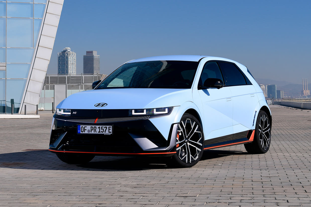 Light blue Hyundai electric vehicle with black and orange accents parked on a paved area with a modern building and city skyline in the background.