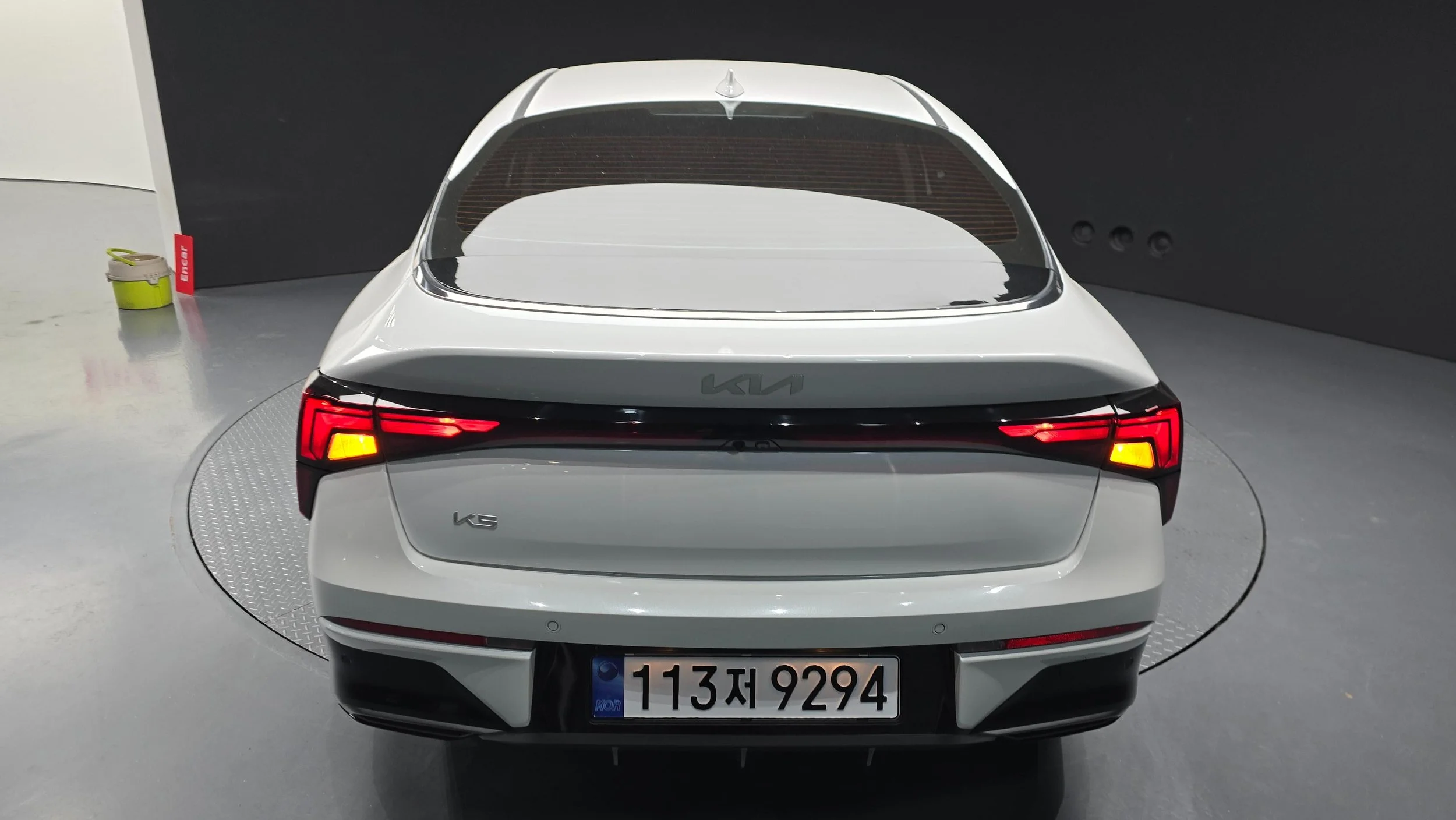 Rear view of a white KIA luxury sedan on a rotating platform inside a showroom, with black trim, LED tail lights, and a Korean license plate.
