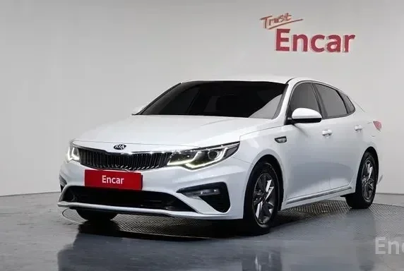 A white Kia sedan parked indoors in front of a white wall with 'Test Encar' signage.
