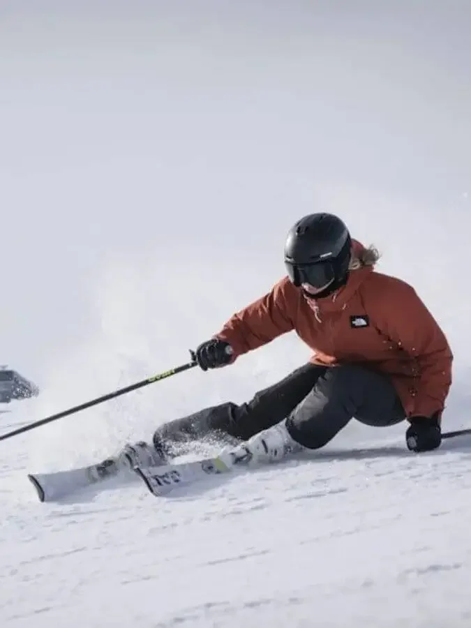 En person i skidåkning placerad på en snöig sluttning, med en orange jacka, svart hjälm och skidutrustning, som gör en svår sväng.