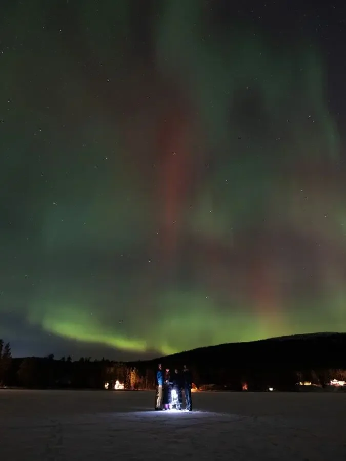 Fyr personer står på ett snötäckt landskap under norrsken i en mörk himmel.