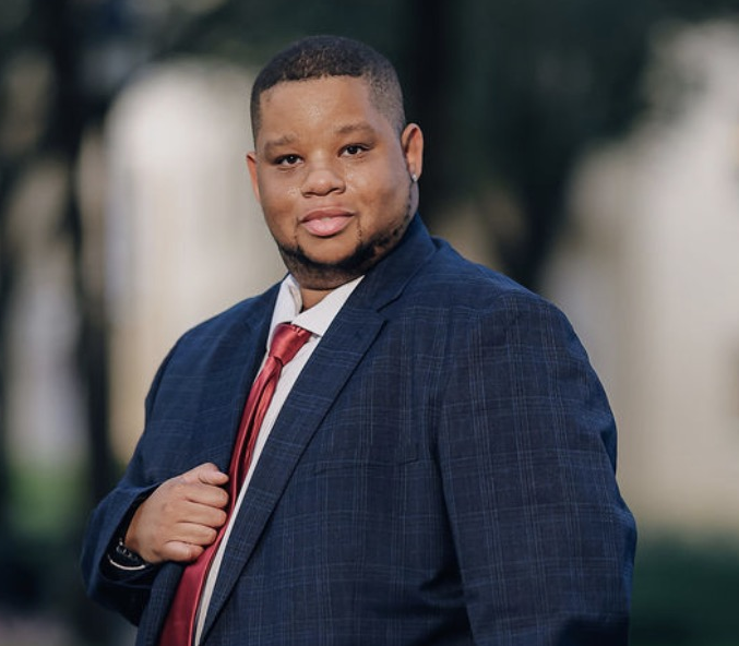 A confident man dressed in a blue plaid suit and red tie, standing outdoors with blurred trees in the background.