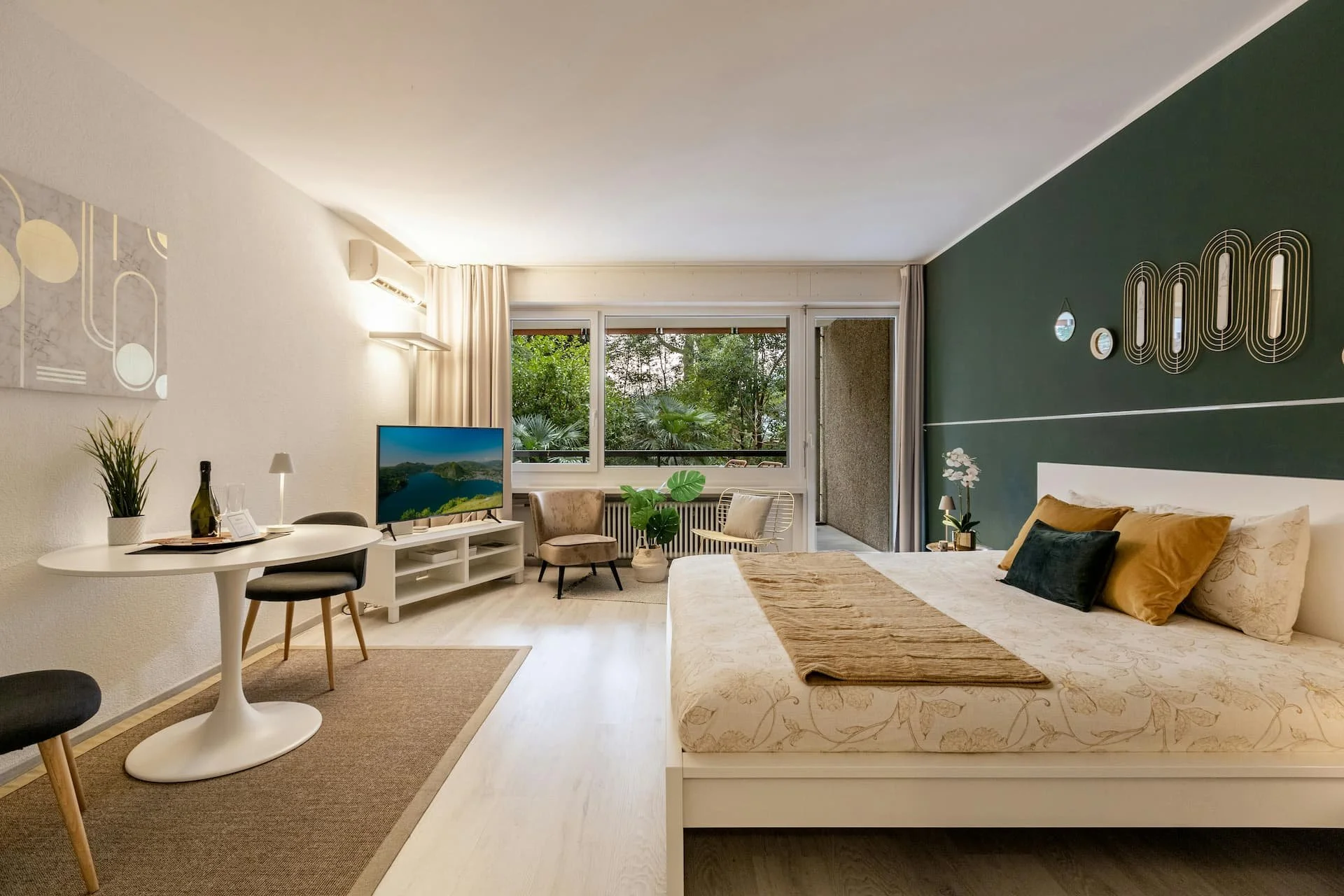 Modern hotel room with a large bed, green accent wall, flat-screen TV, small sitting area, and balcony with trees outside.