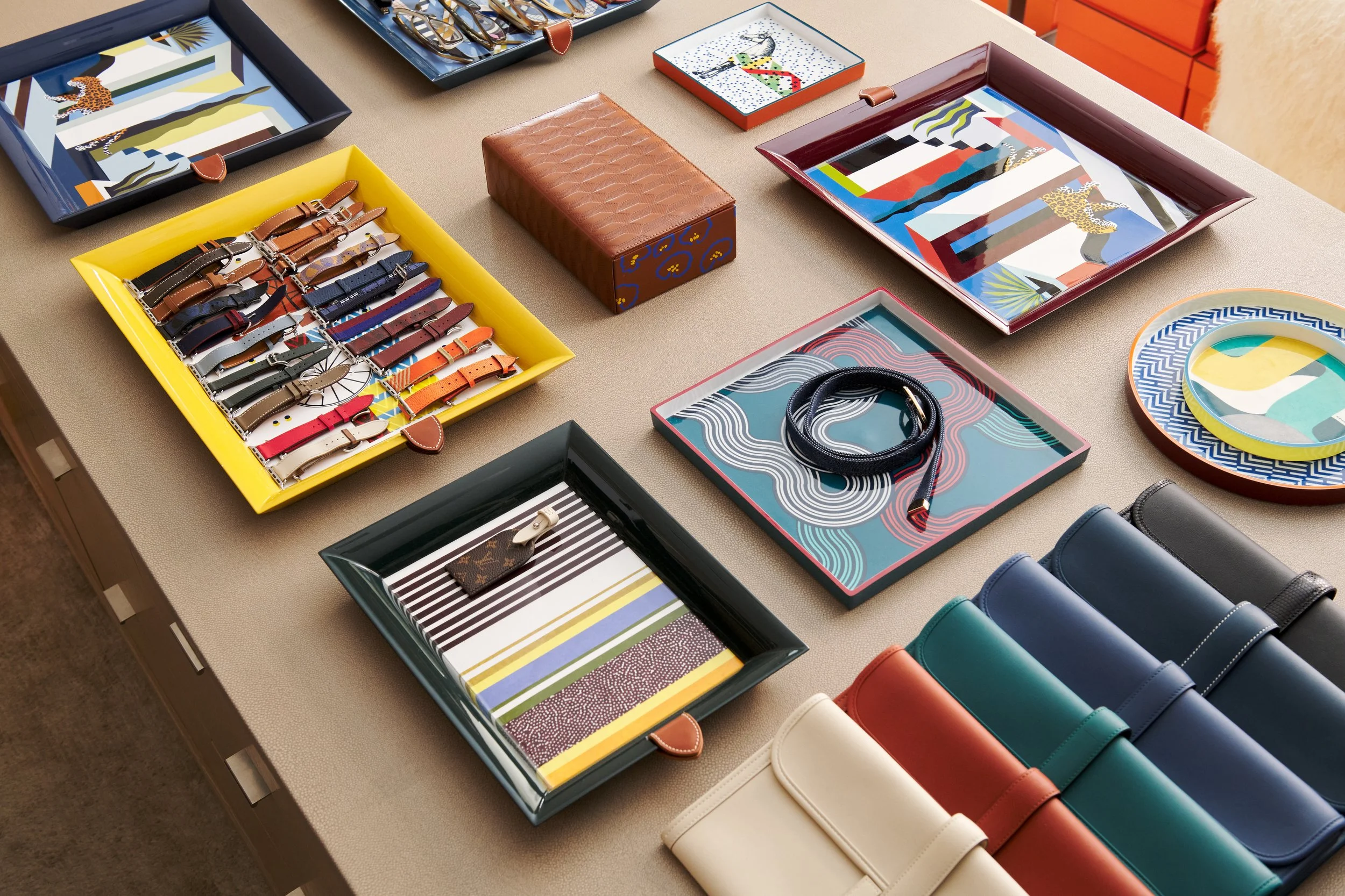Display table showcasing various fashion accessories including colorful watches in a yellow box, designer keychain in a black box, belts in a tray, and wallets in different colors, surrounded by decorative plates and trays.
