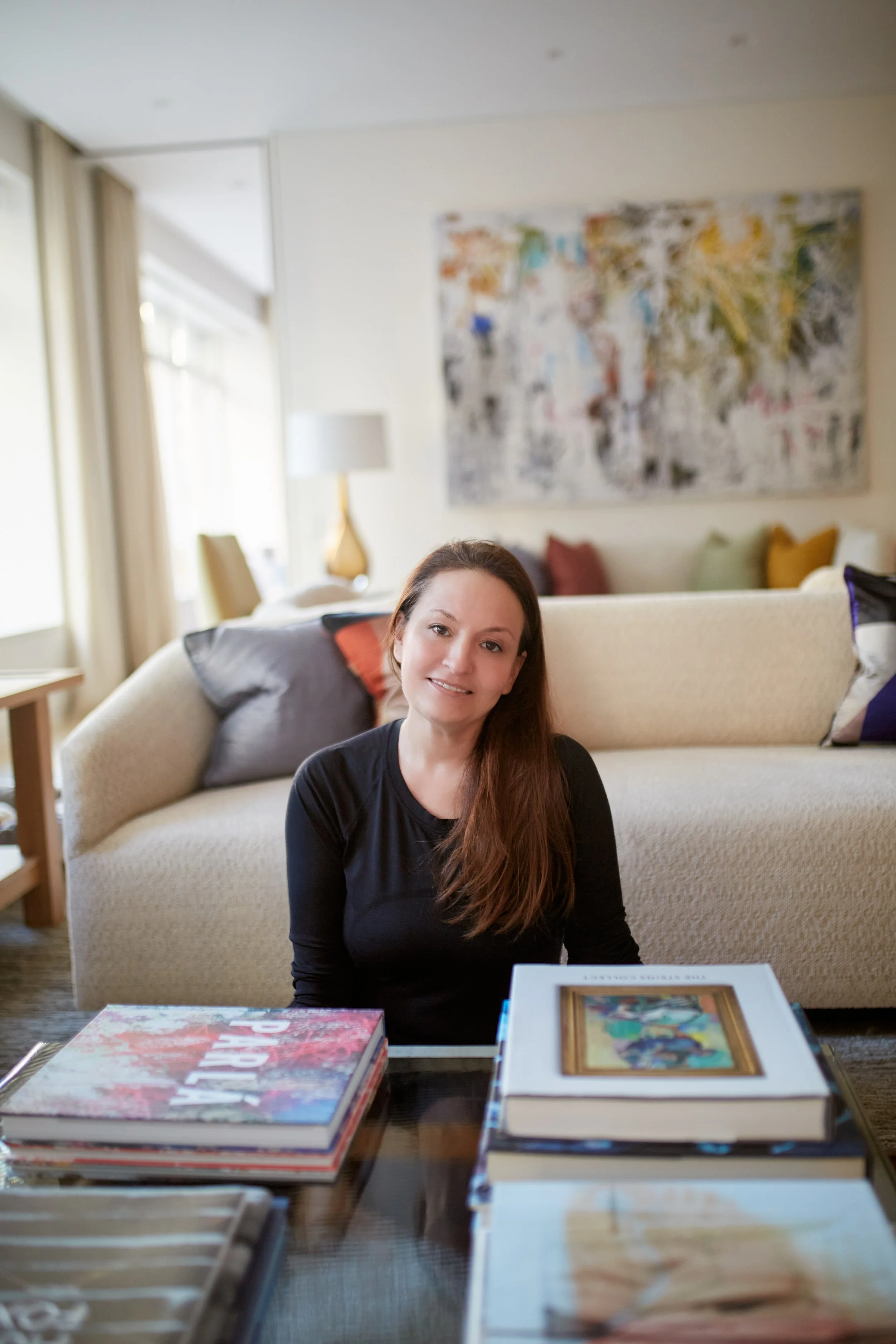 A woman with brown hair and a black shirt sitting on the floor in a living room, smiling at the camera with a stack of art books on the glass coffee table in front of her.