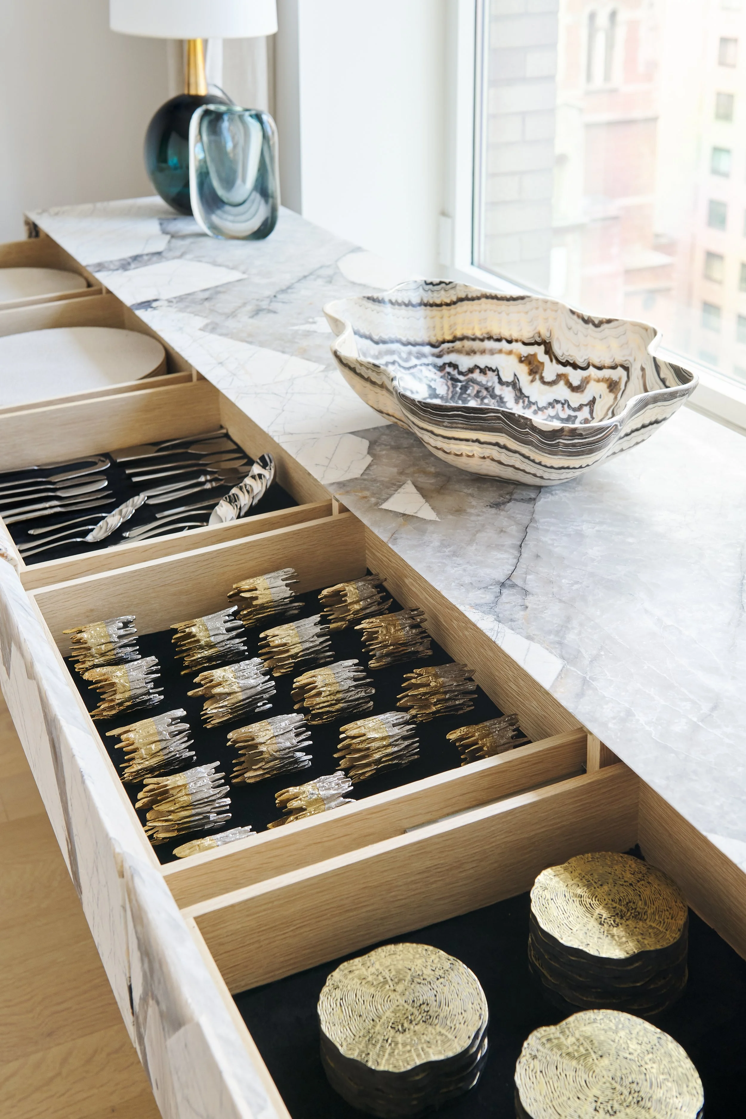A marble and wood dresser with jewelry, tableware, and a decorative bowl, near a window with an urban view.