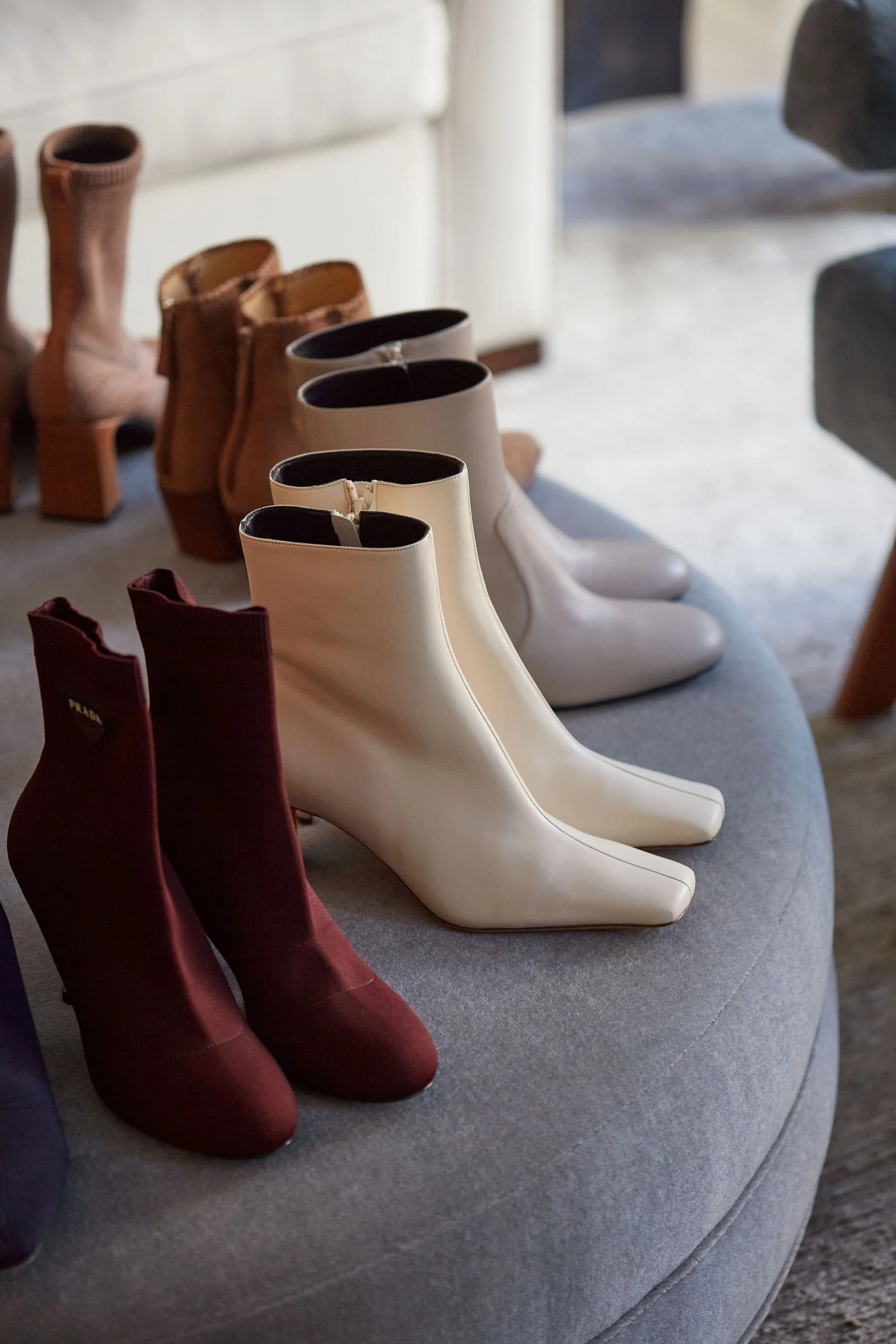 Collection of women's footwear, including maroon ankle boots, white pointed-toe ankle boots, and beige heeled boots, displayed on a grey upholstered bench.