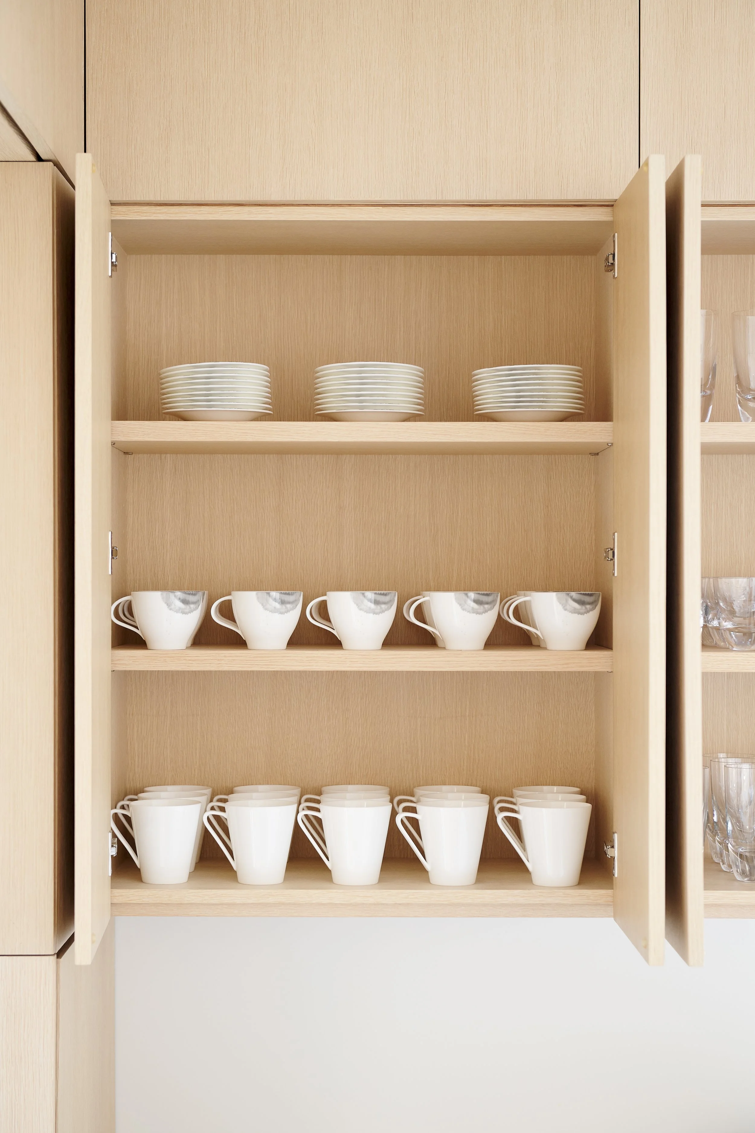 Open kitchen cabinet with three shelves containing white dishes, cups, and glasses.