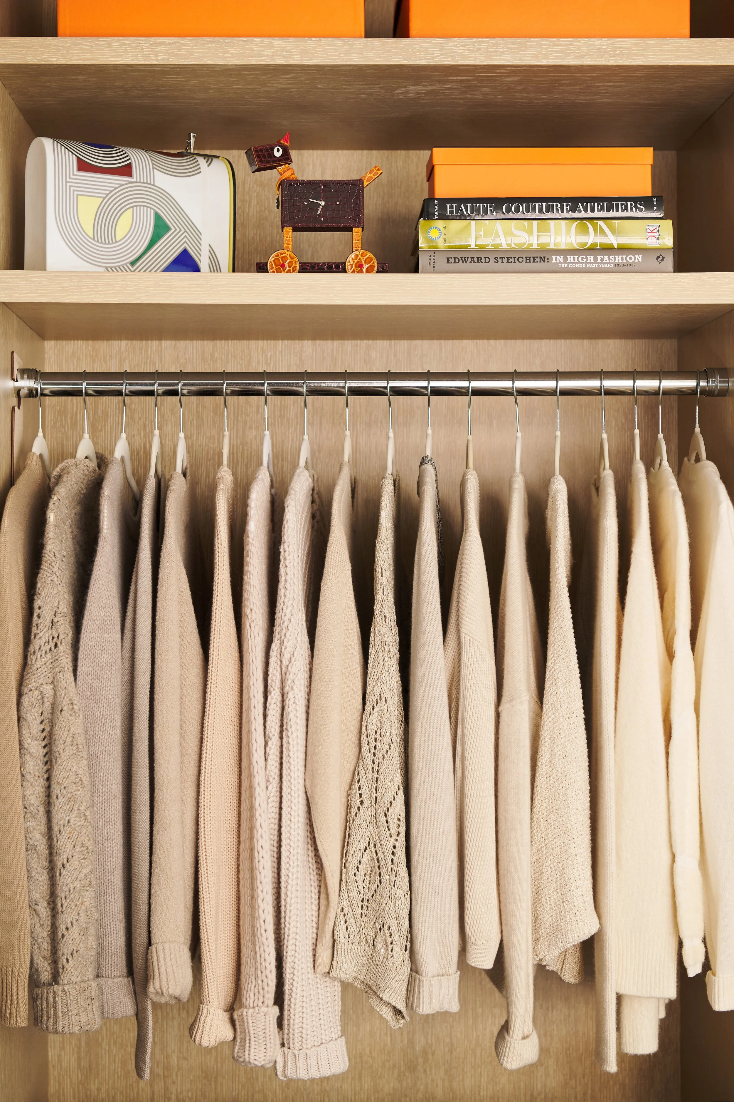 A closet with a row of beige and cream sweaters and shirts hanging on a metal rod, with three shelves above containing books, a decorative box, and a small giraffe-shaped figure.