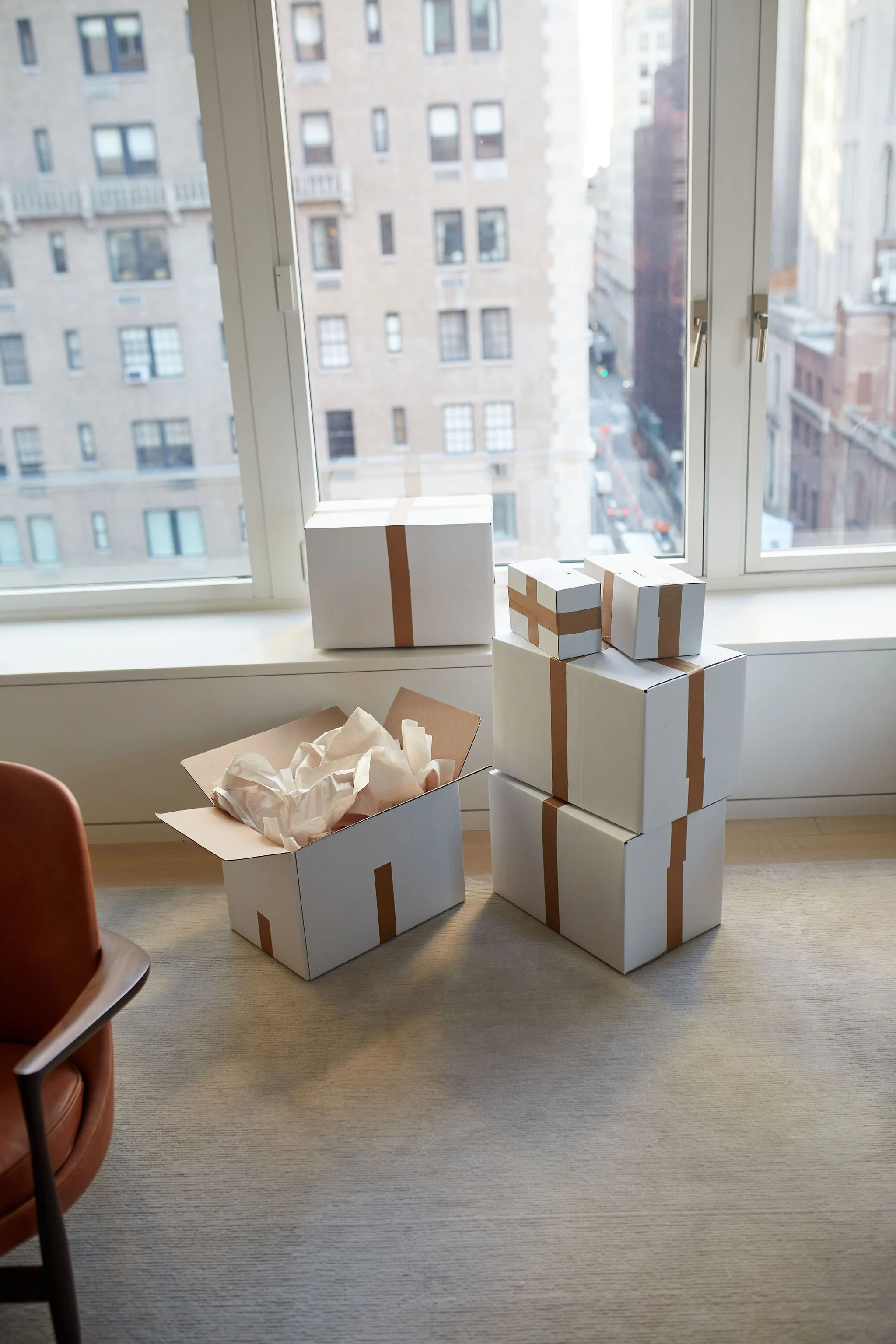 Several white boxes with brown tape, some open with tissue paper, are stacked near a large window overlooking a city street.