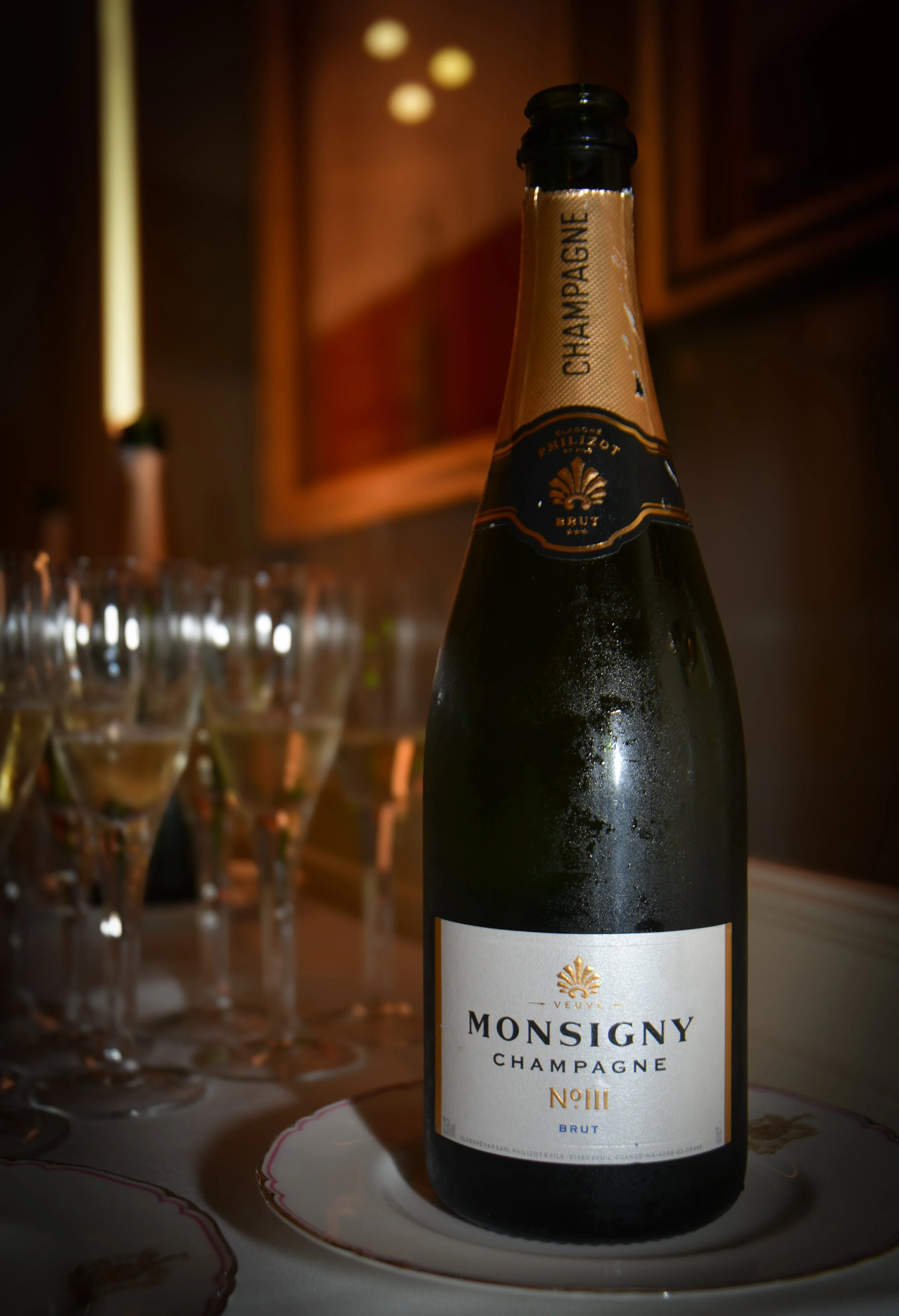 Bottle of Monigny Champagne Brut with condensation, placed on a plate in front of a row of champagne glasses on a table, in a dimly lit room.