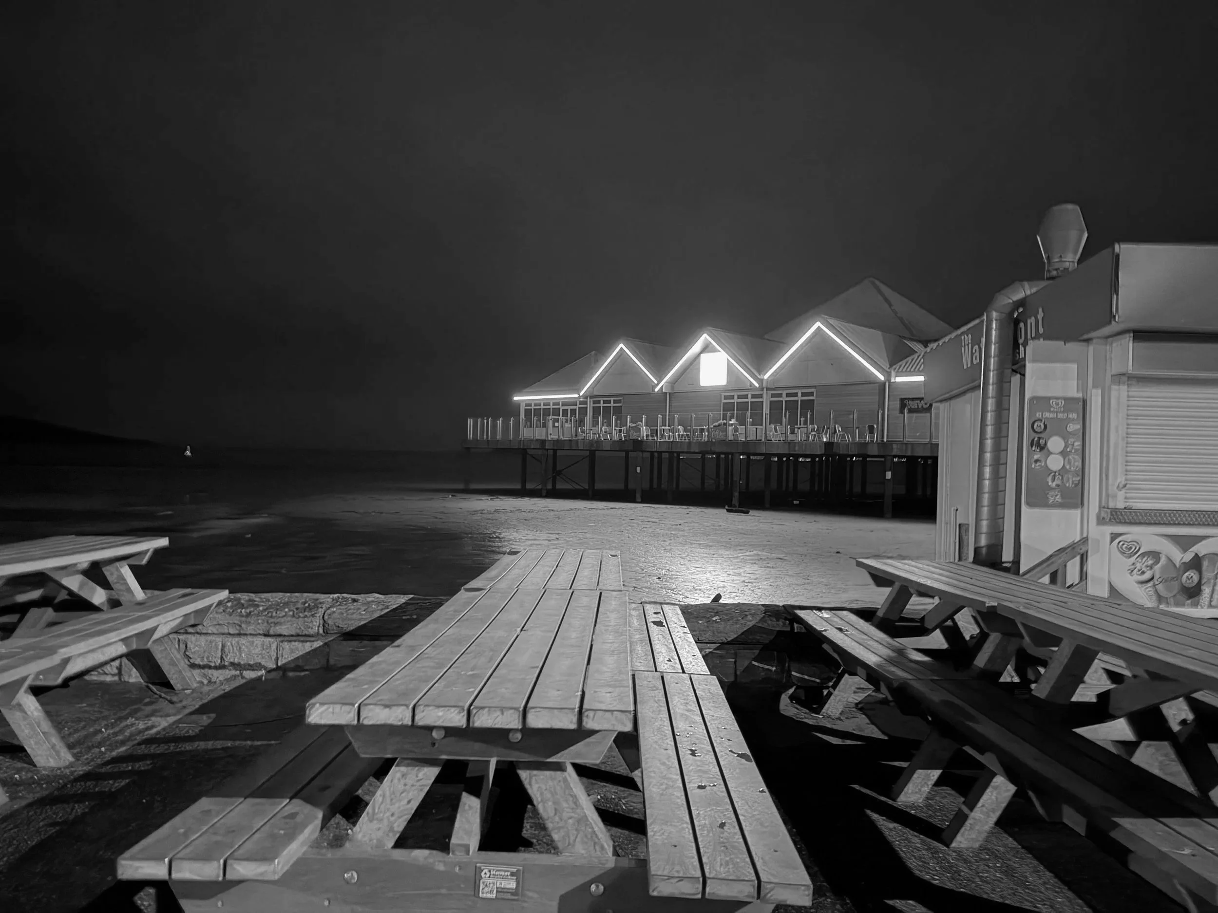 Empty wooden picnic tables on the beach at night, with a shadowy pier and illuminated building with neon lights in the background.