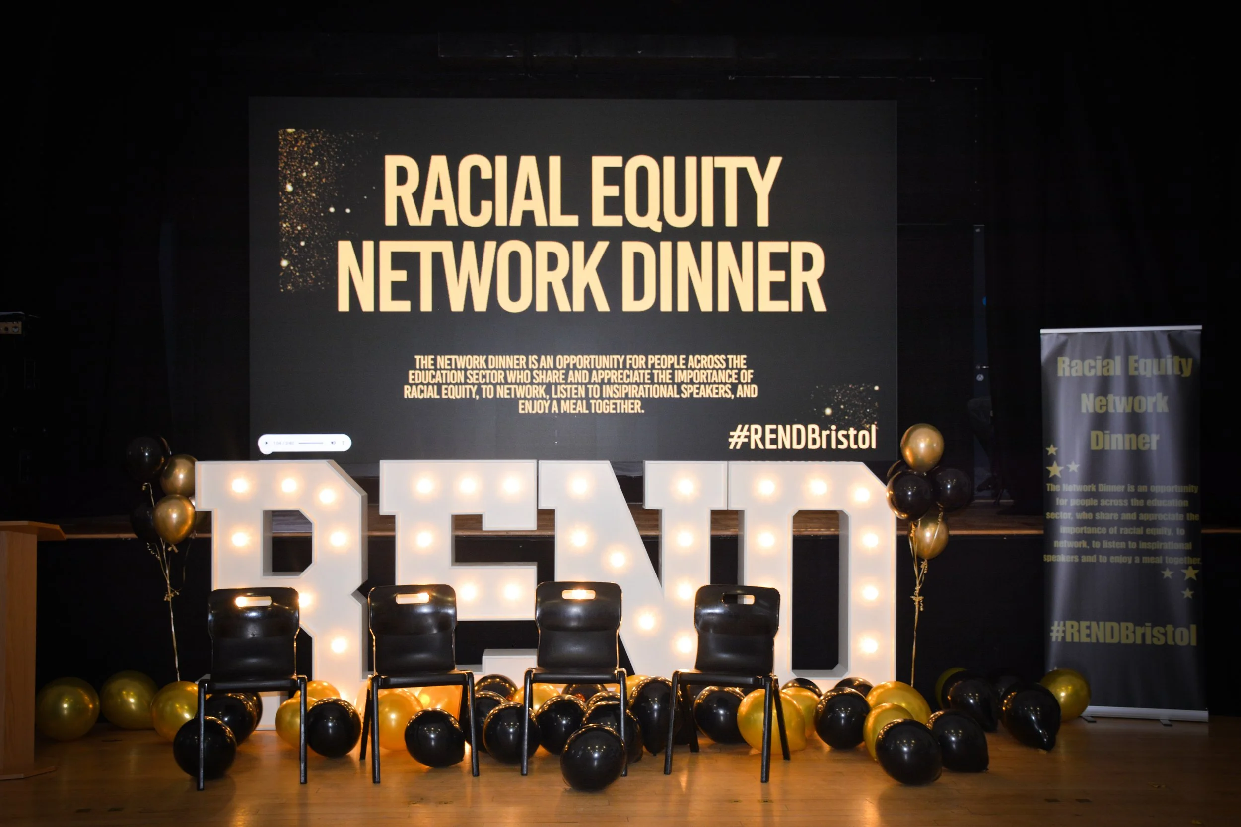 Decorative setup with illuminated letters spelling 'BEY' on stage, black and gold balloons, large screen displaying 'Racial Equity Network Dinner' with information, and banner with event details in the background.