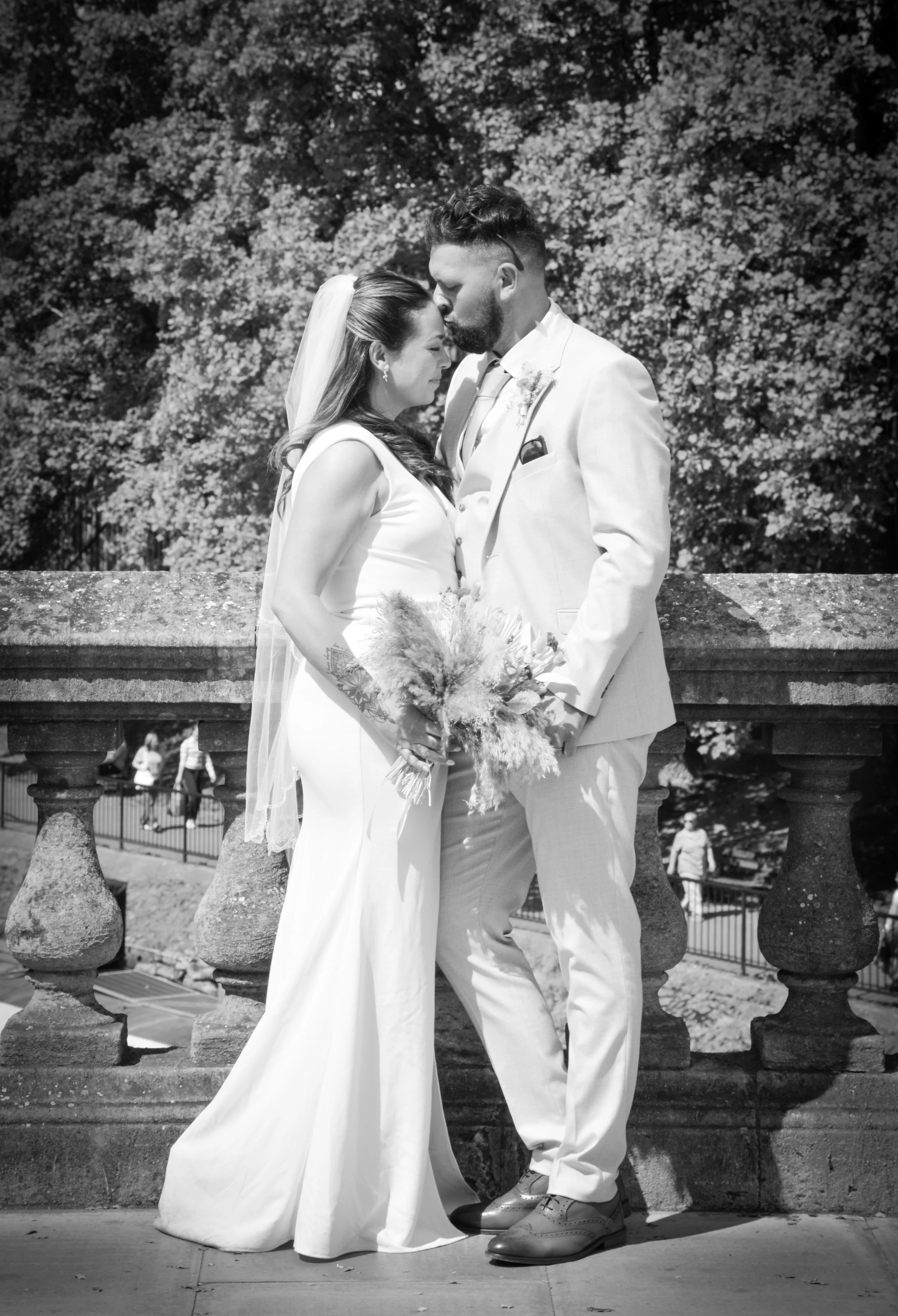 Black and white photo of a bride and groom standing close, forehead to forehead, in front of a stone railing, with trees in the background.
