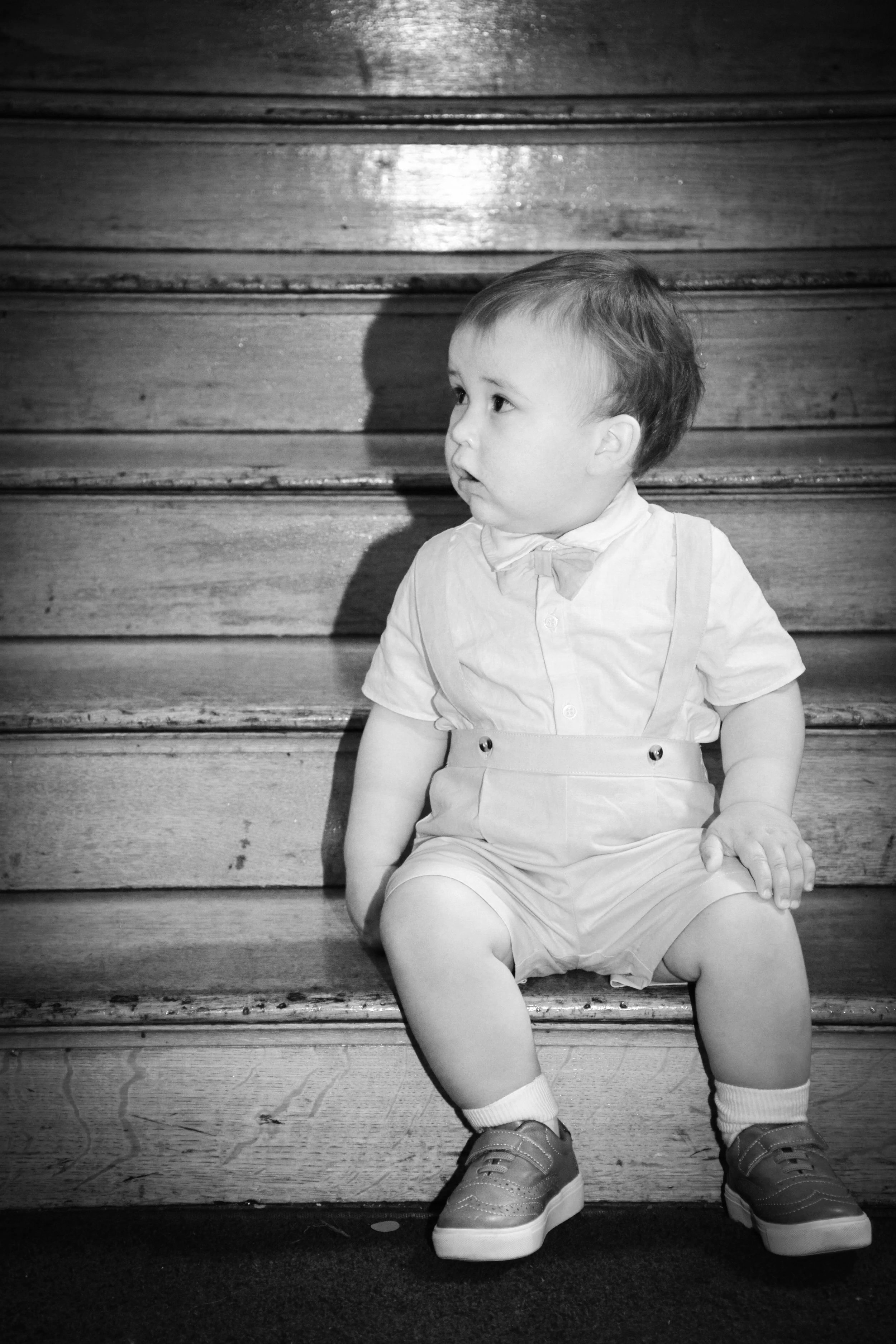 A young boy sitting on wooden stairs looking to the side, wearing a light-colored shirt, shorts, sneakers, and a bow tie.