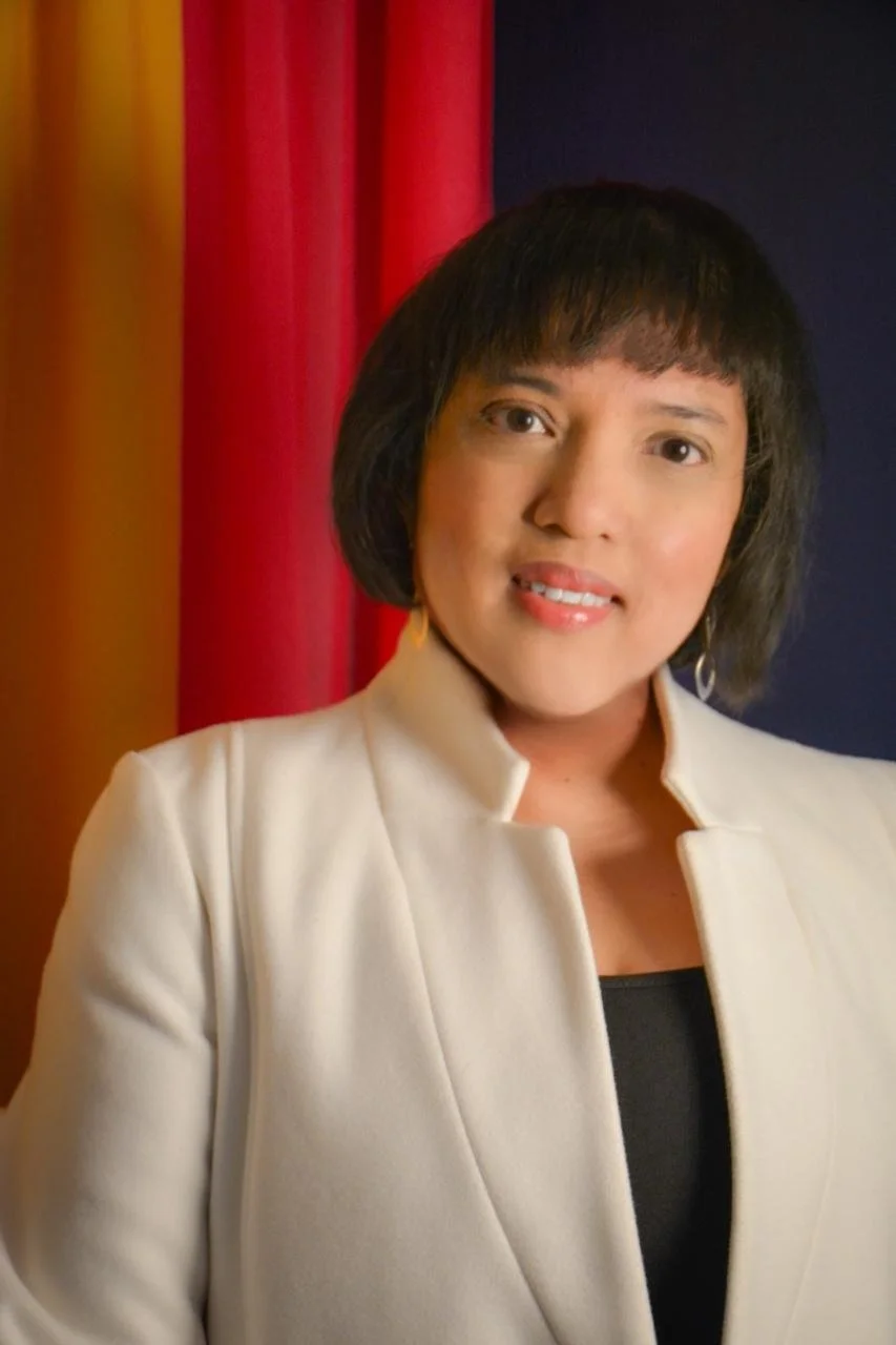 A woman with short black hair wearing a cream-colored blazer and black top standing in front of red and dark blue curtains.