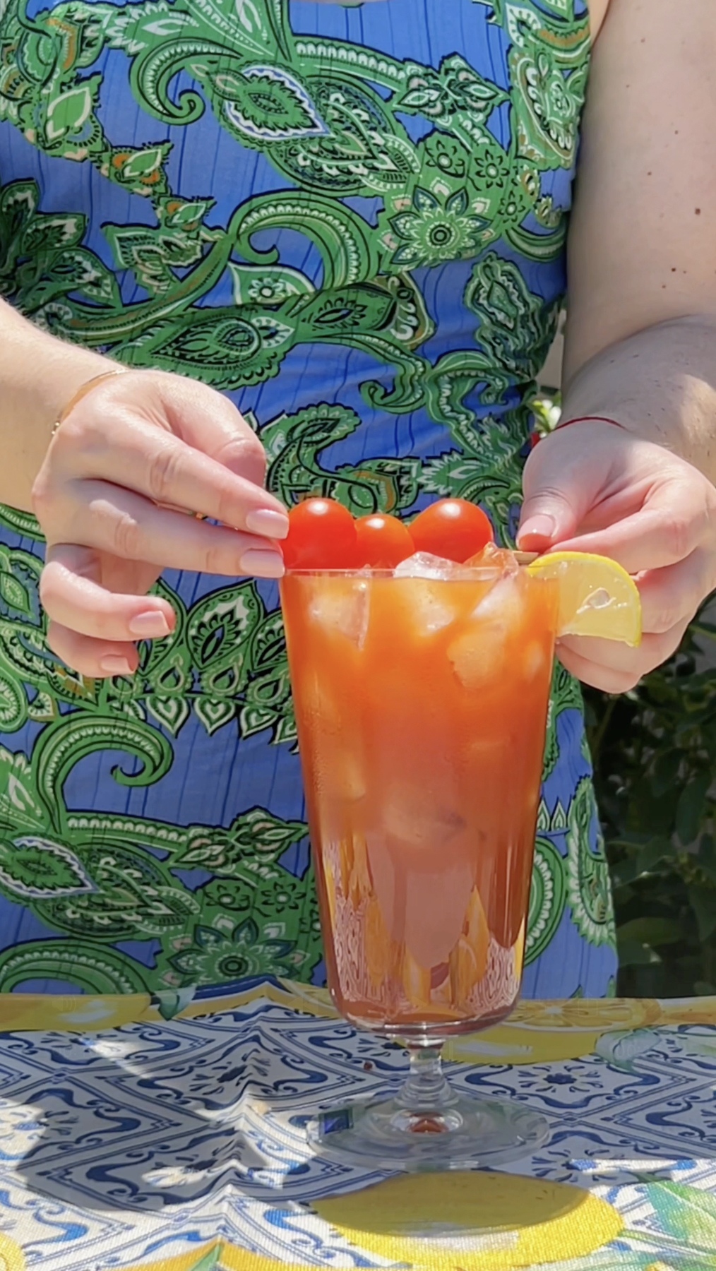 A person in a blue and green patterned shirt is holding a tall glass of tomato cocktail garnished with cherry tomatoes and a lemon wedge. The tablecloth below has blue and yellow decorative patterns.