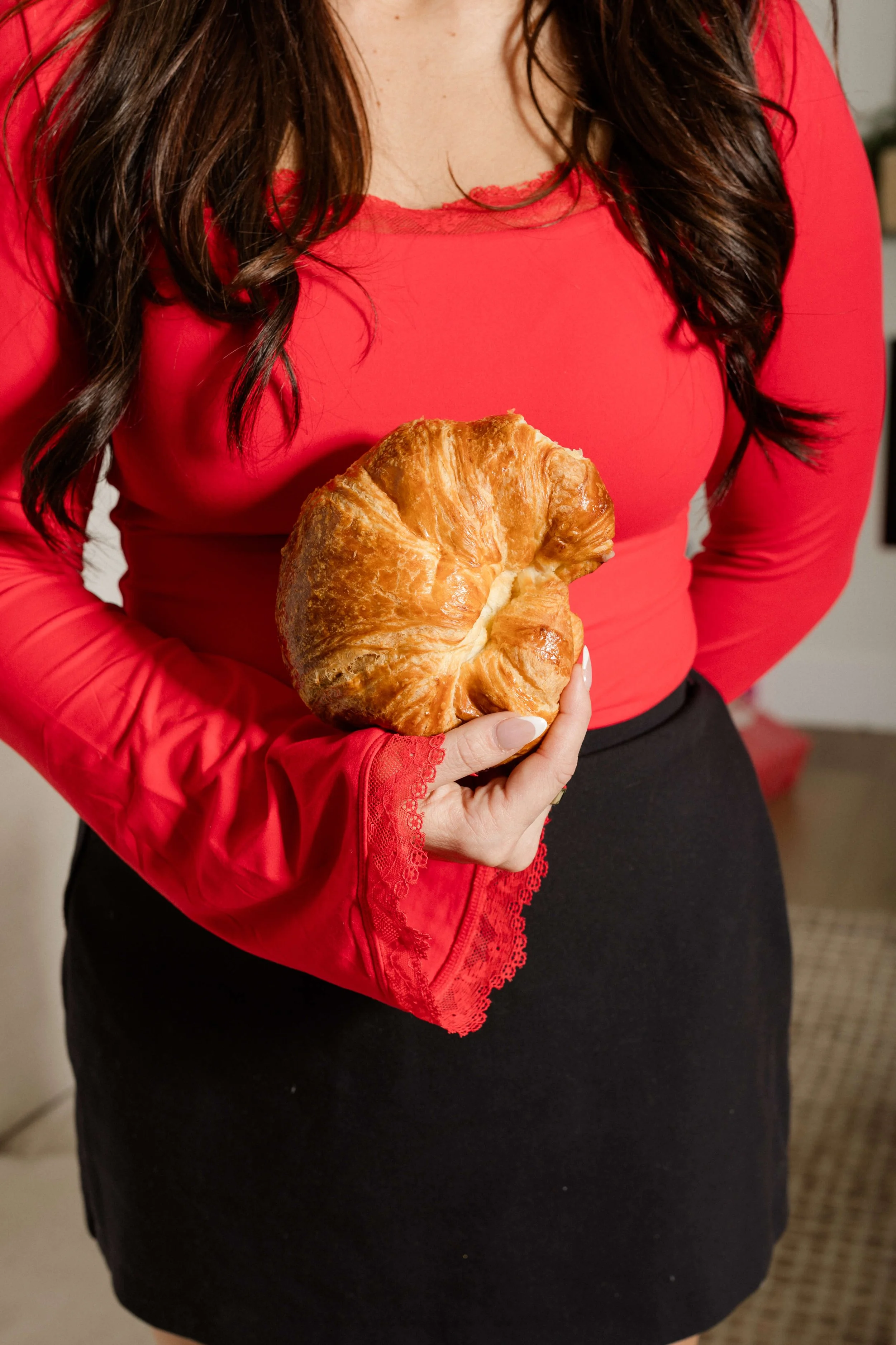 A woman with dark brown hair wearing a red top with lace trim holding a croissant in her right hand.