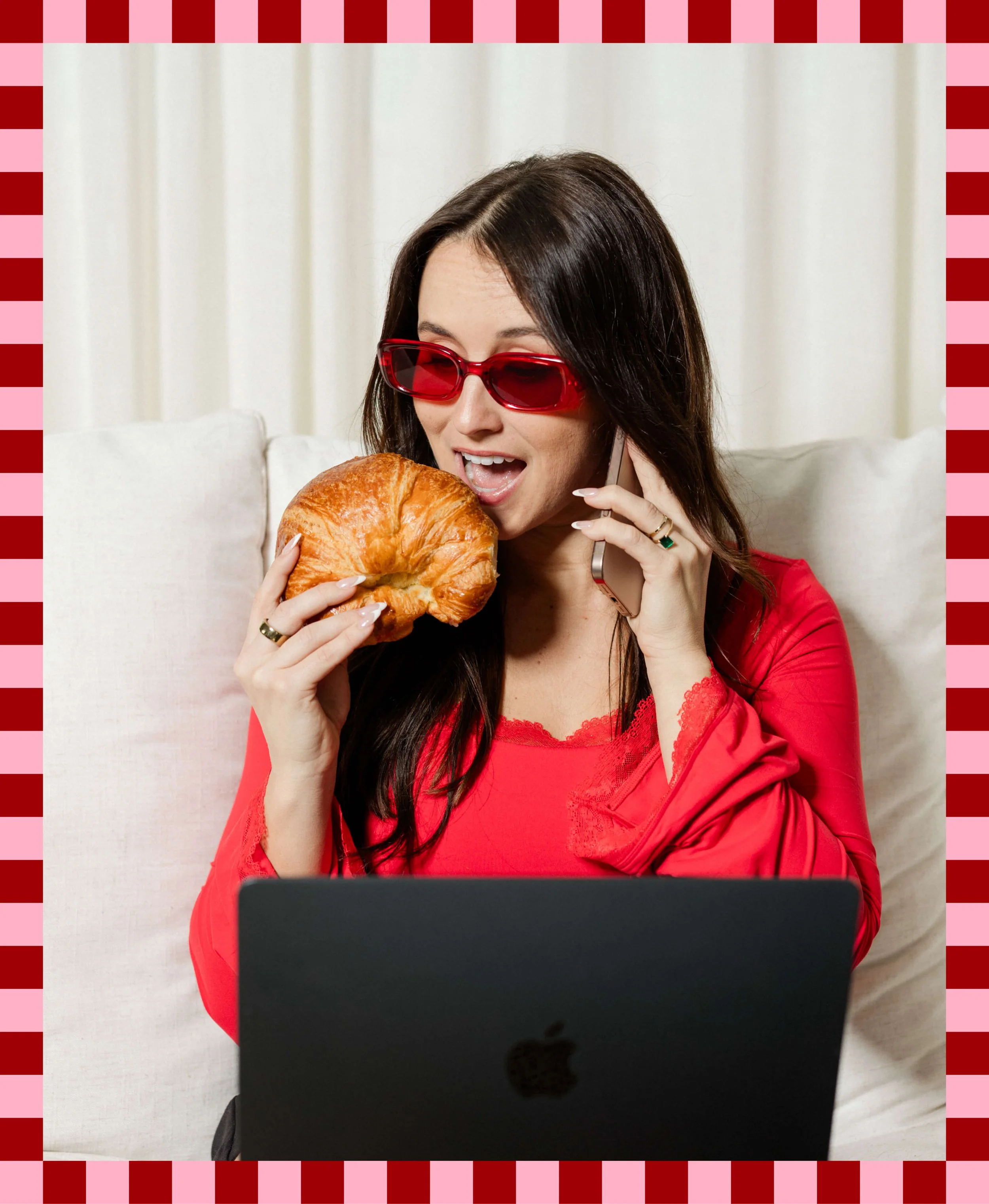 Woman with long dark hair wearing red sunglasses and red blouse, talking on a cellphone while holding a croissant in front of her face, sitting on a beige couch with white curtains in the background, and a laptop in front of her.