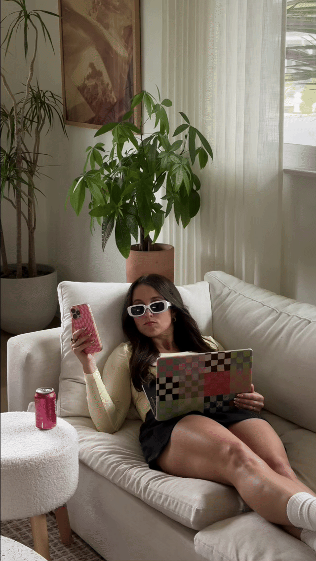 A woman with long dark hair, wearing sunglasses, a cream-colored sweater, and a black skirt, is reclining on a white sofa. She is holding a pink phone in her right hand and a colorful checkered laptop in her lap. There is a pink soda can on a small round white table beside her. The background features large potted plants, a framed artwork, and sheer curtains next to a window.