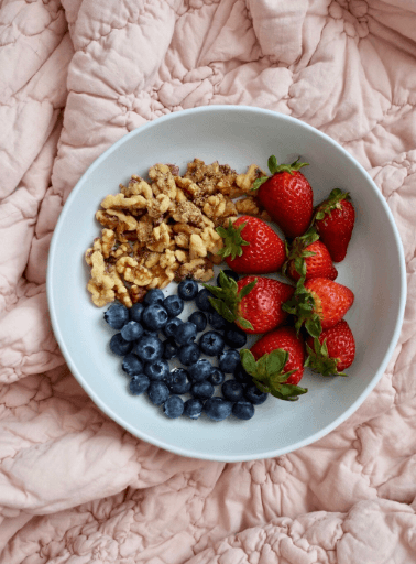 Bowl of fresh strawberries, blueberries, and granola on a pink quilted blanket.