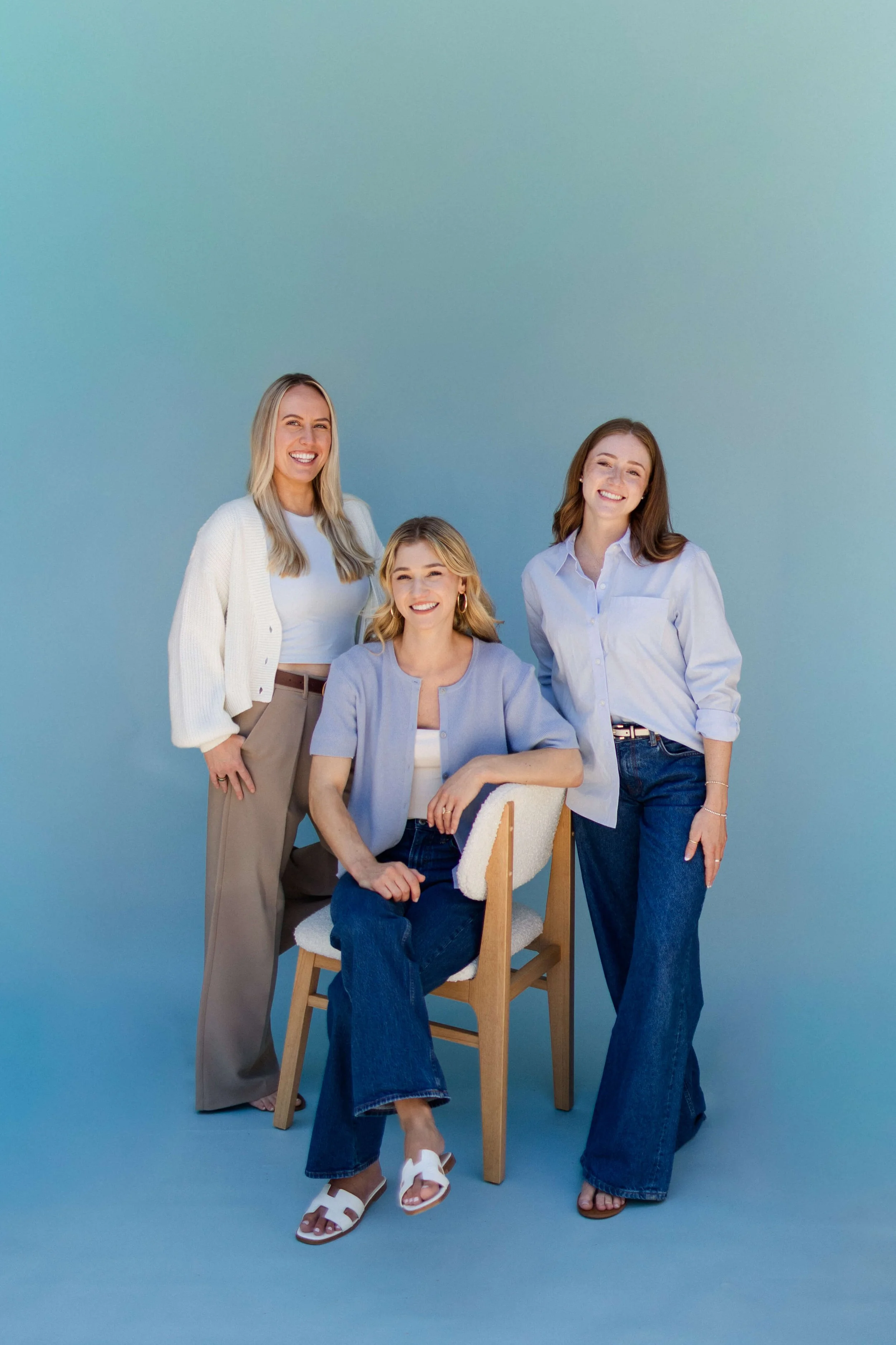 Four women smiling, standing and sitting against a solid blue background. One woman is seated in a chair with a cardigan over a top, another is standing in a white blouse and jeans, a third woman in a white top and beige pants, and the fourth in a white shirt and dark jeans.