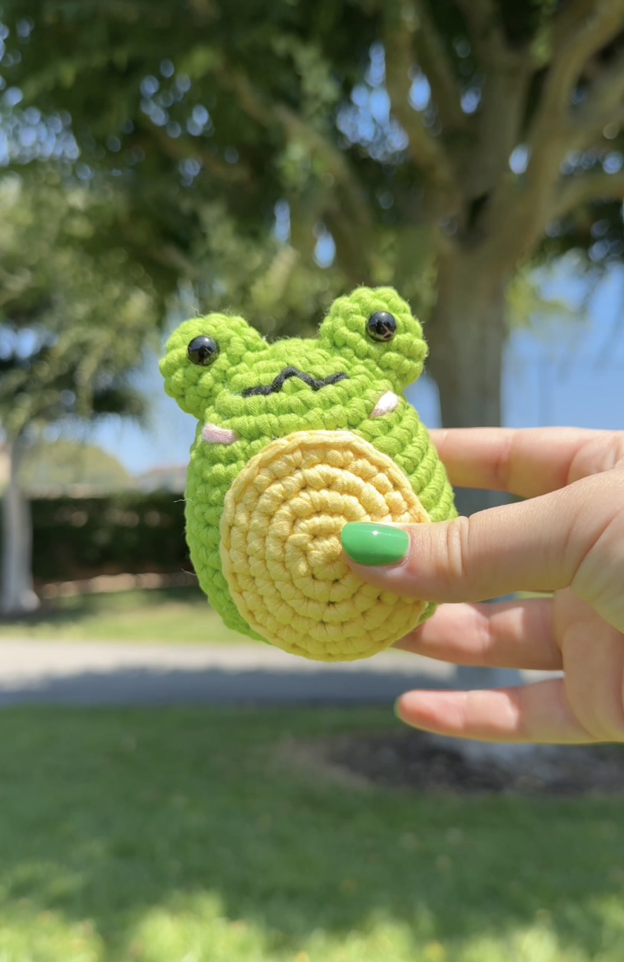 Close-up of a hand holding a plush toy of a green frog with black eyes and a yellow belly outdoors with trees and a blue sky in the background.