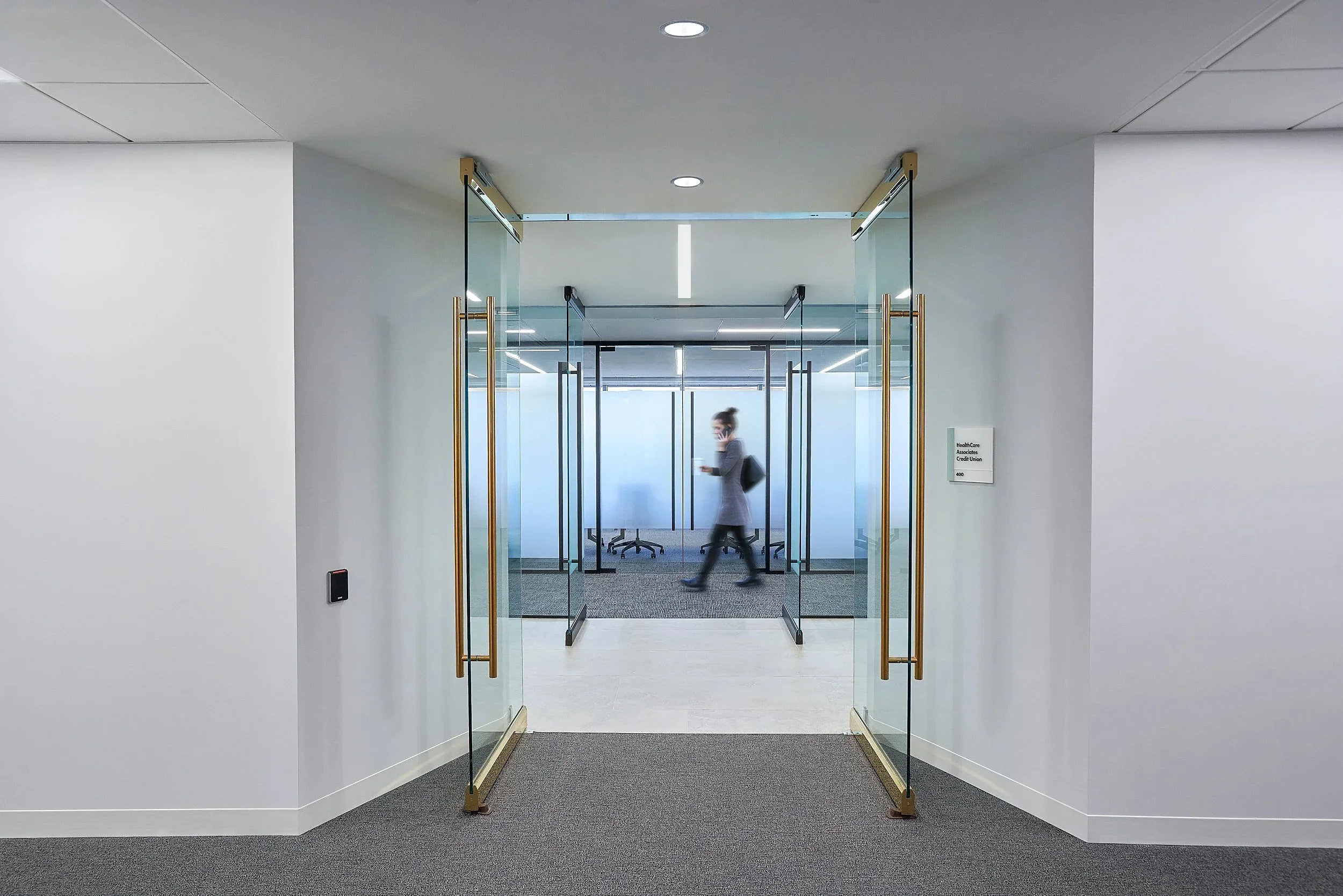 Interior view of a modern office space with glass doors and a woman walking while talking on her phone.