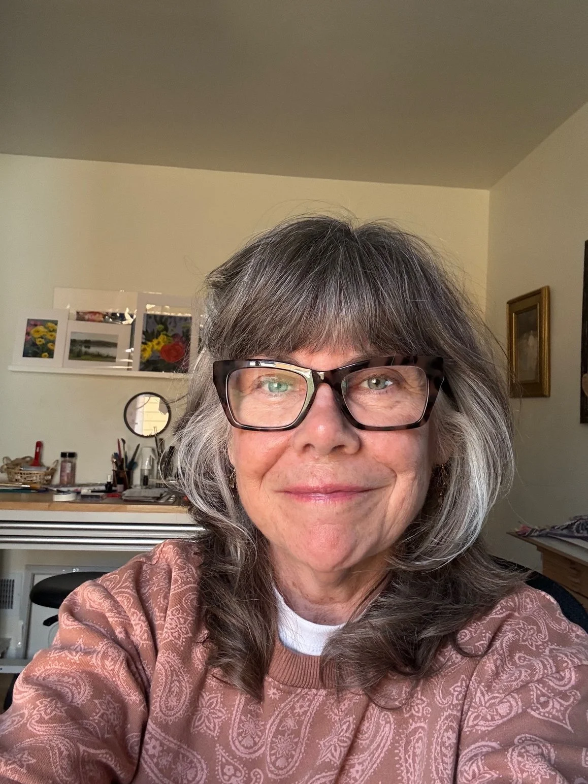 A woman with shoulder-length gray hair, wearing glasses and a patterned brown top, smiling in a room with art supplies and framed pictures on the wall.