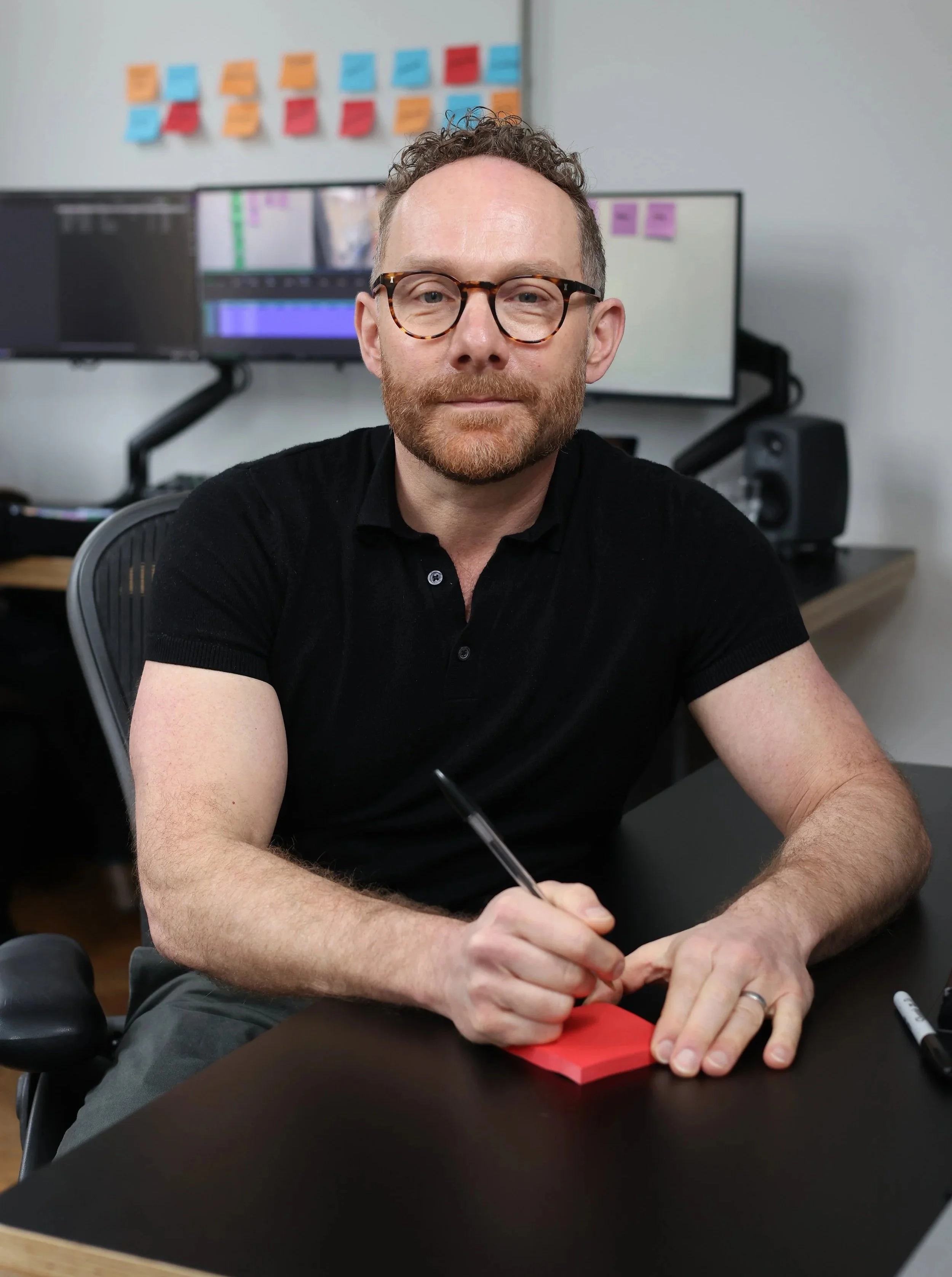A man with curly hair and glasses sits at a desk with a pen and red sticky notes, in an office with dual computer monitors and colorful sticky notes on the wall behind him.