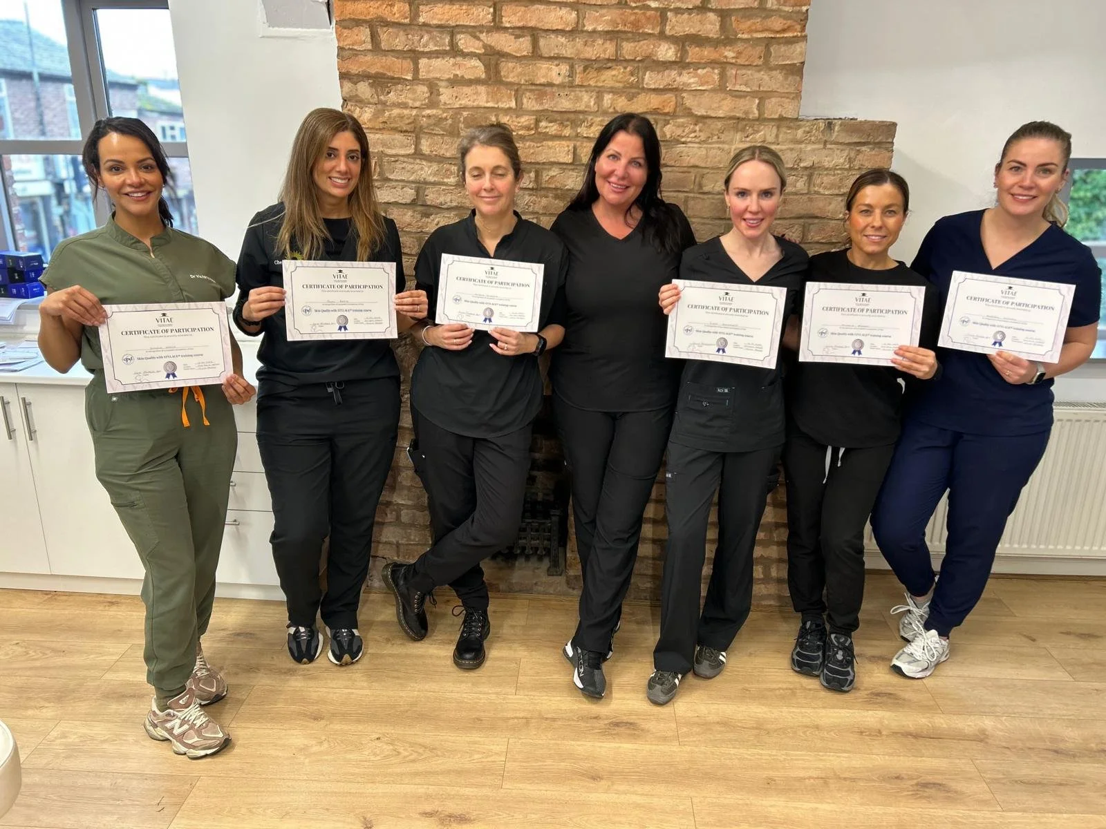 A group of seven women standing together indoors, holding certificates of participation. They are dressed in medical or professional uniforms and smiling at the camera with a brick wall behind them.