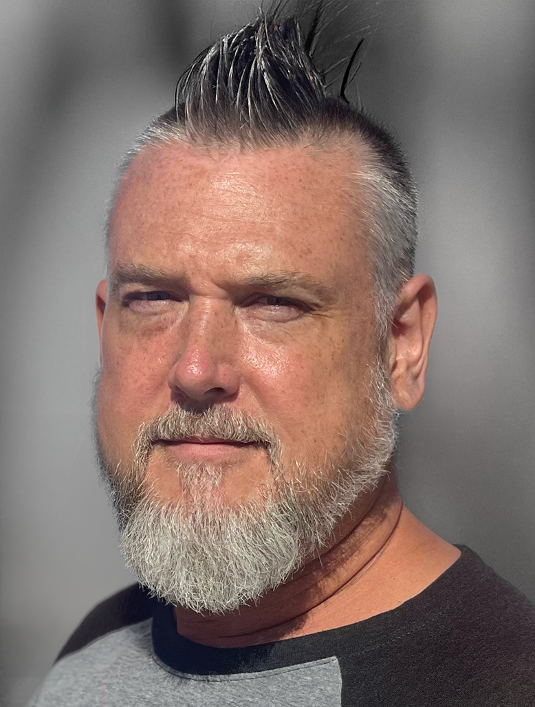 A middle-aged man with a spiked hairstyle, white beard, and wearing a black and gray shirt, standing indoors with a neutral gray background.